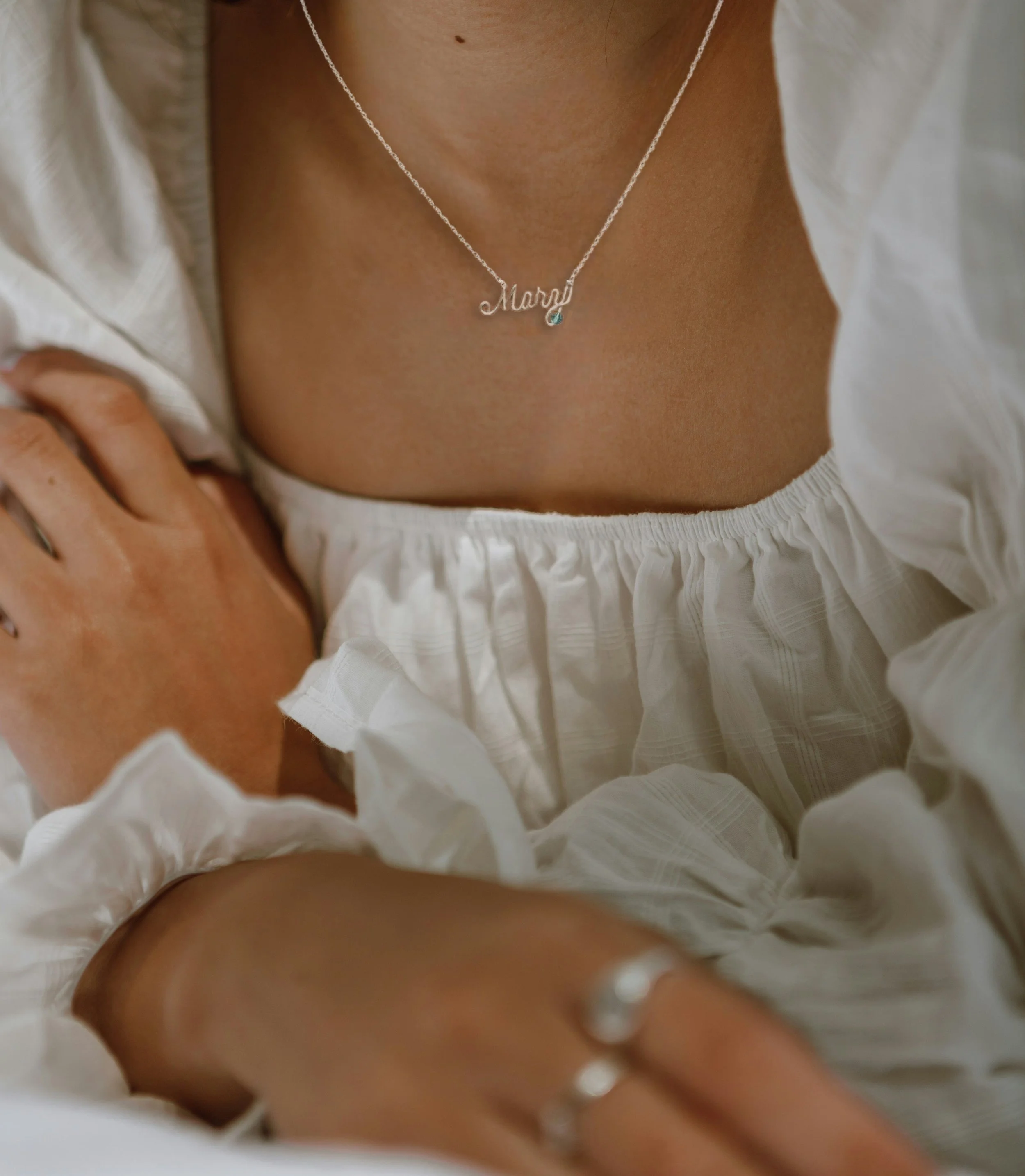 A close-up of a woman wearing a silver necklace with the name 'Mary' and a small blue gemstone, white puffy top, with rings on her fingers.