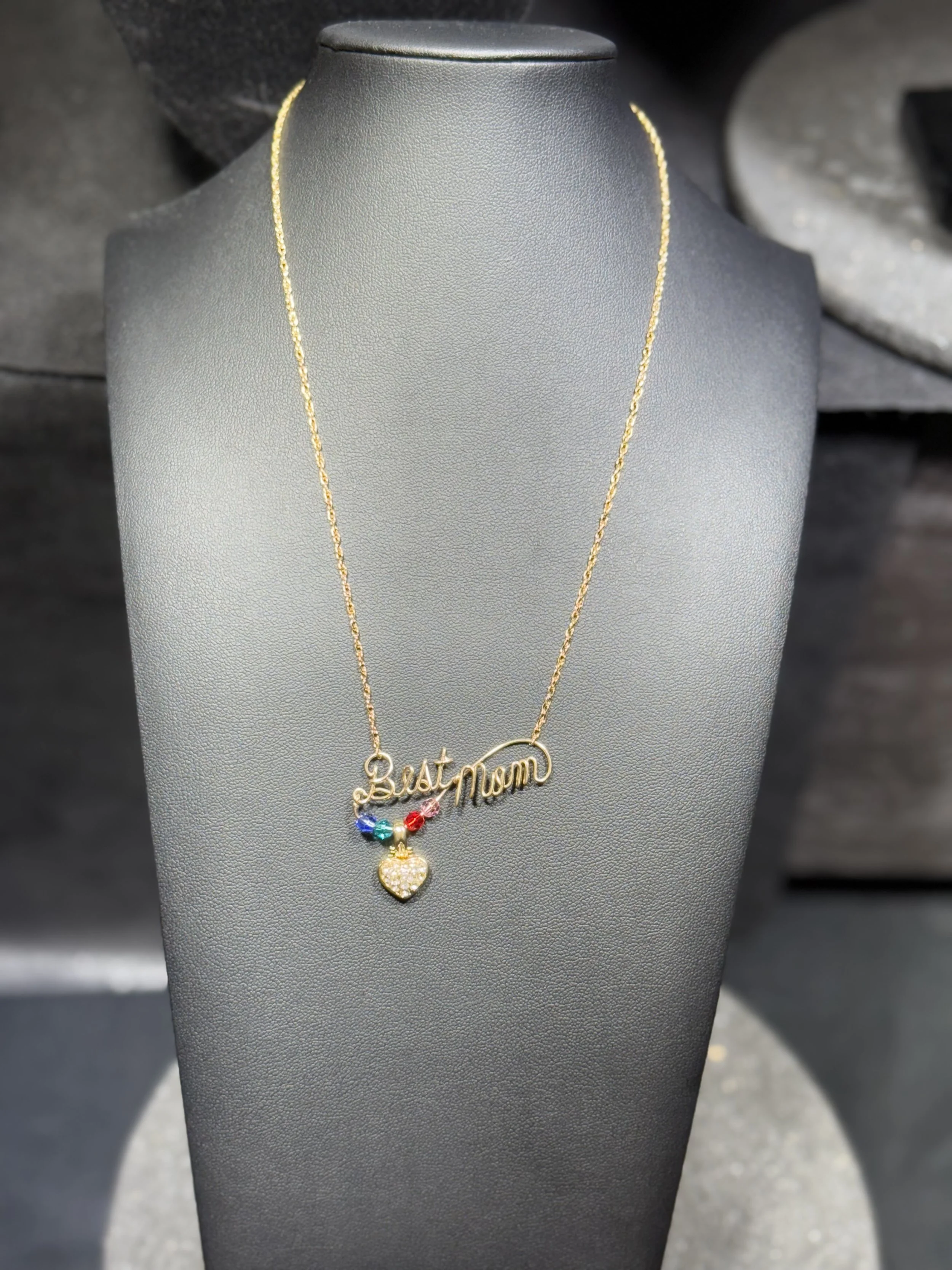 Gold necklace with a heart-shaped pendant and multicolored beads, with a charm that reads 'Best Mom' in cursive script, displayed on a black jewelry stand.