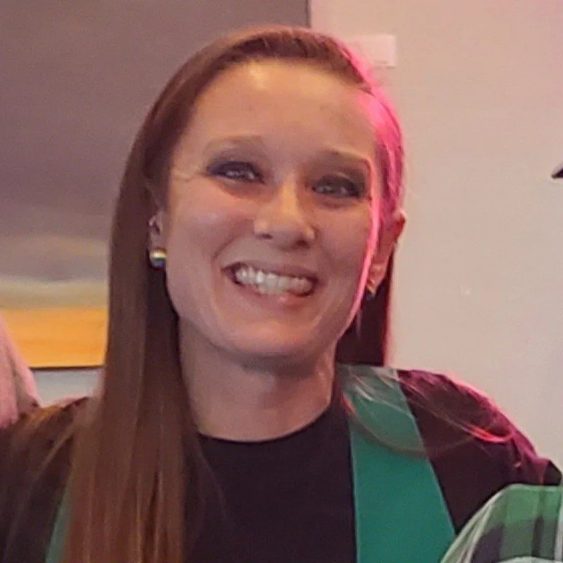 A woman smiling with straight brown hair, wearing earrings and a dark top, in an indoor setting with a painted wall and a framed picture in the background.