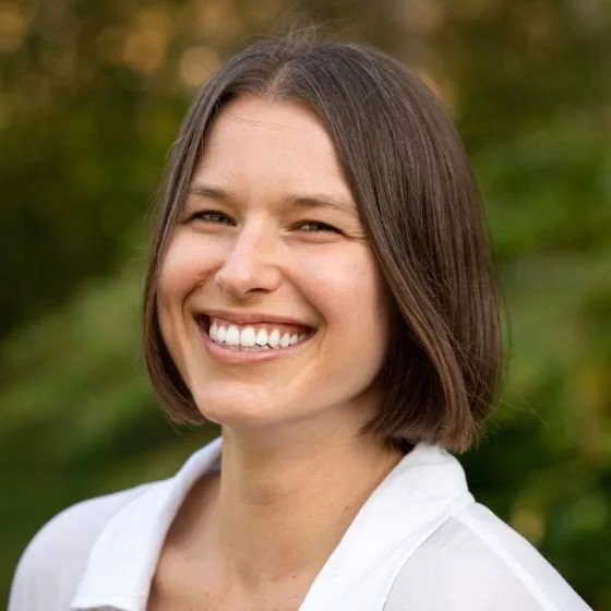 A woman with short brown hair smiling outdoors with a blurred green background.