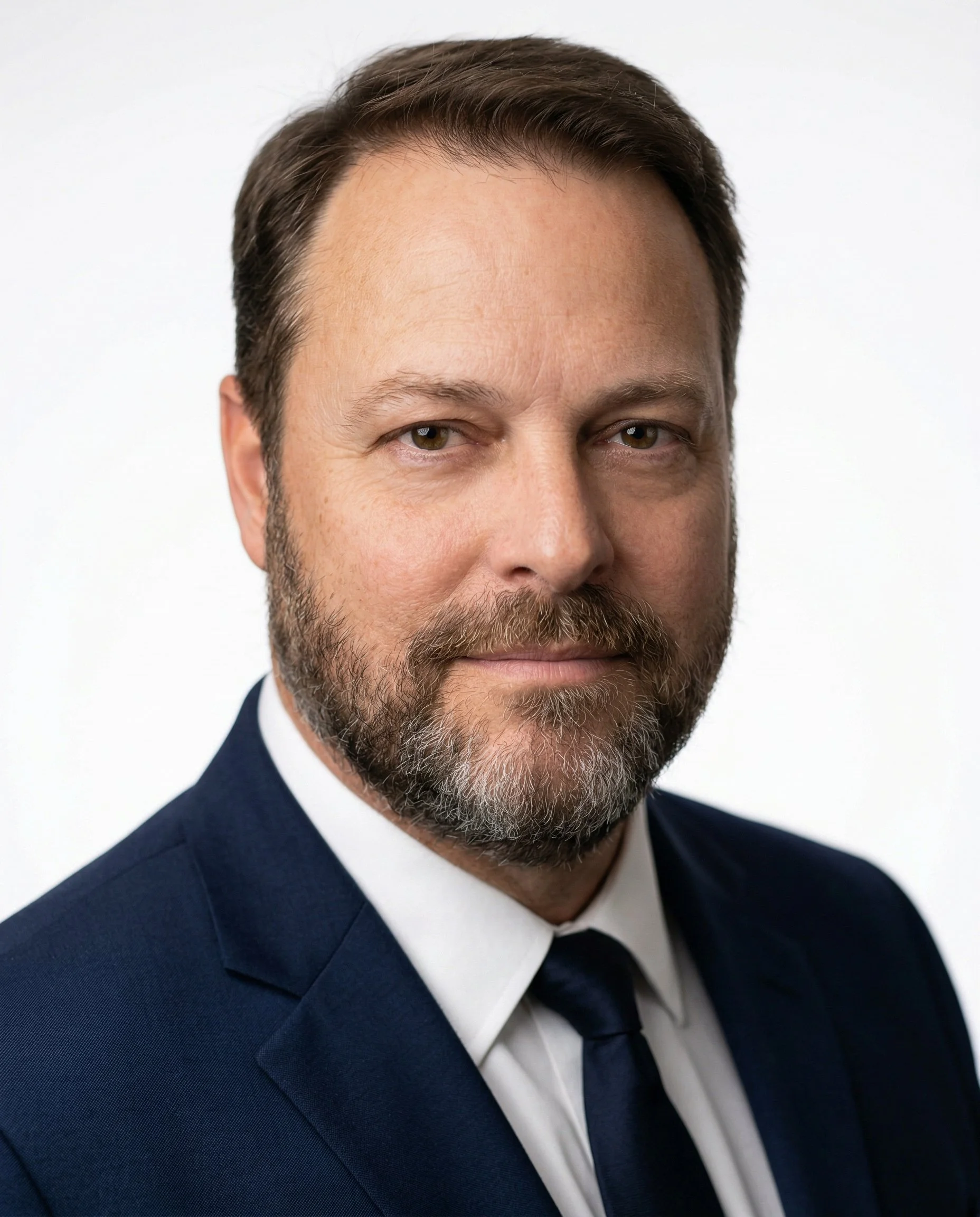 Portrait of a man with light skin, short brown hair, and a beard. He is wearing a black suit and white shirt and black tie, smiling slightly, against a plain beige wall background.