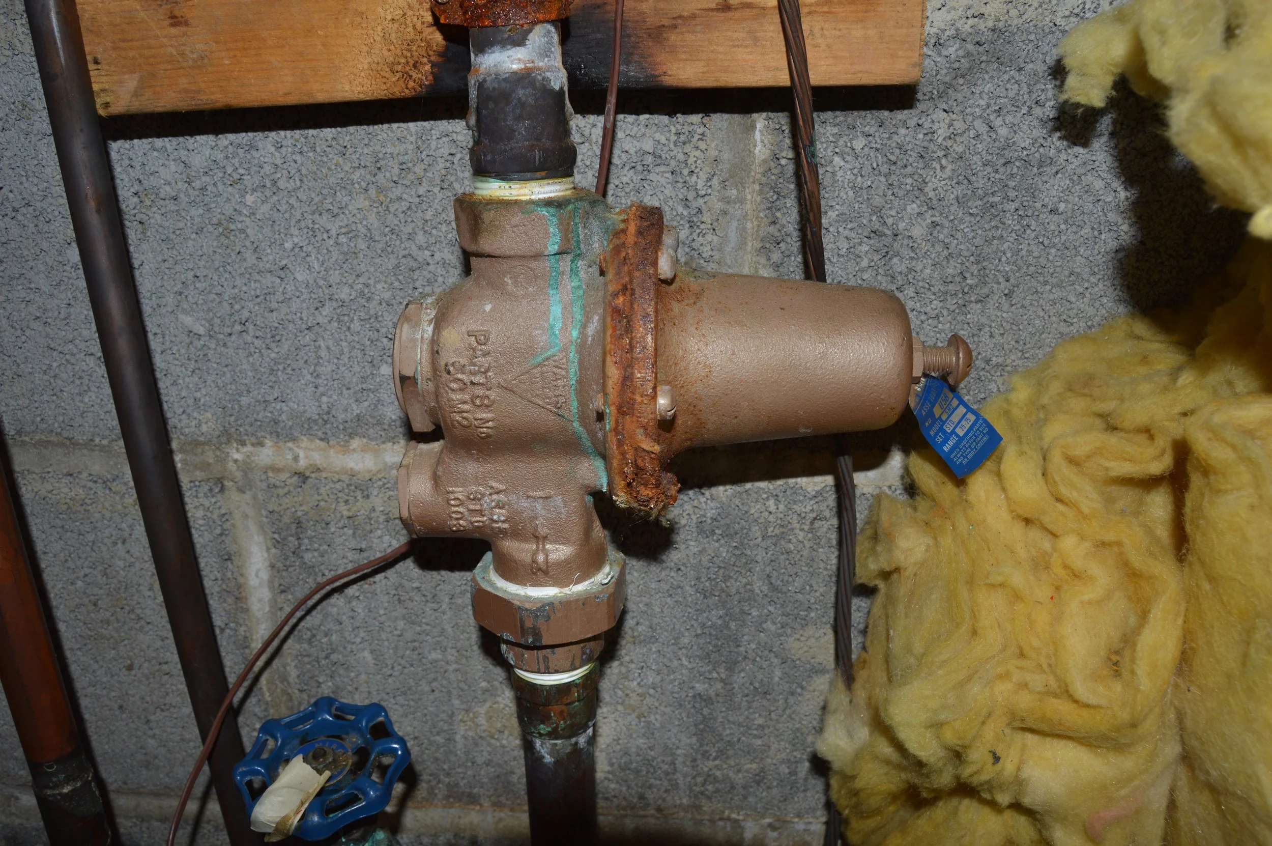 A rusty water valve connected to a pipe with a wall and yellow insulation in the background.