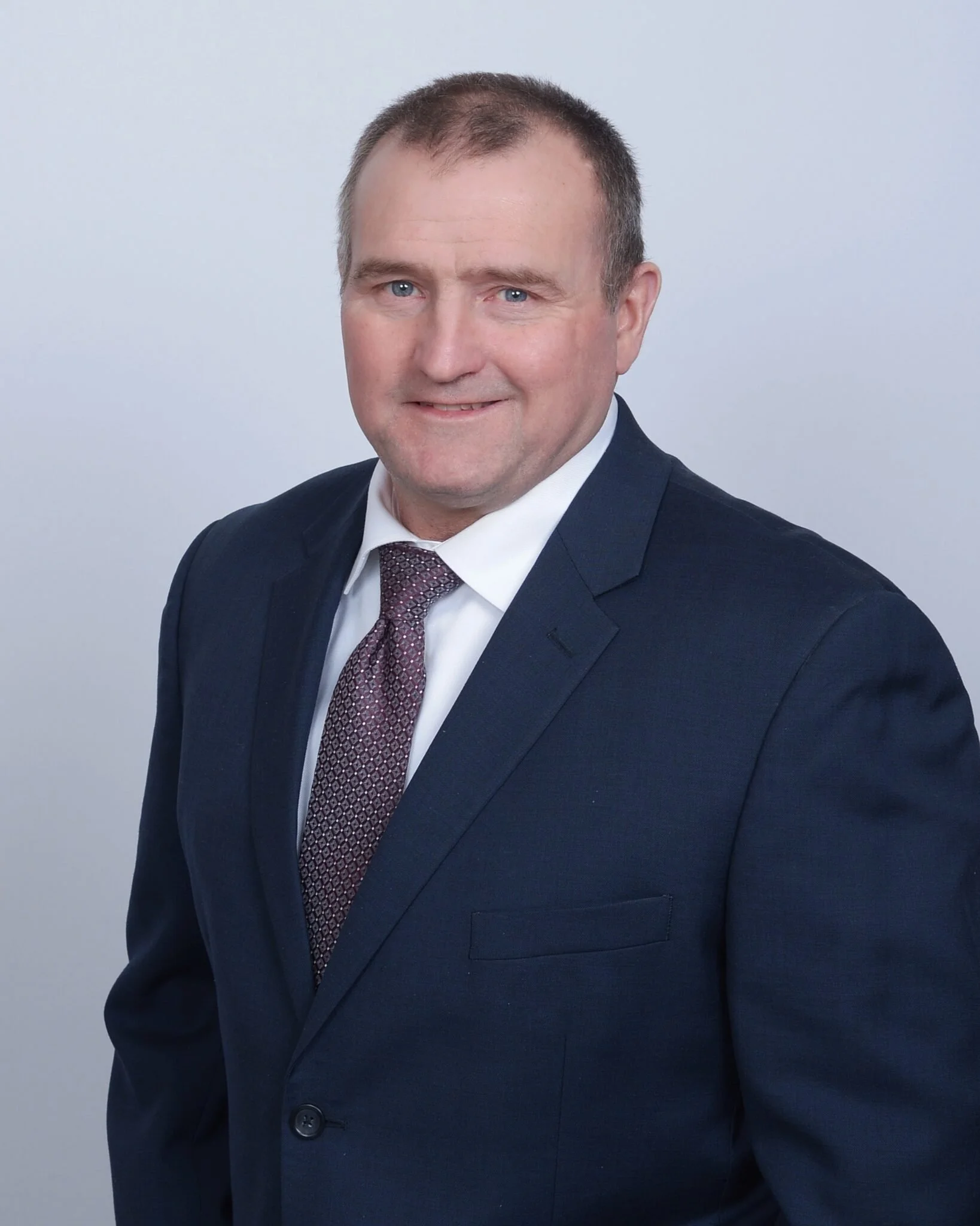 A man wearing a dark suit, white shirt, and patterned tie, smiling at the camera against a light gray background.