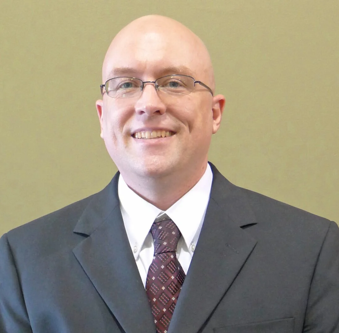 A man in a black suit, white shirt, and patterned tie, wearing glasses, smiling at the camera against a plain, light-colored background.