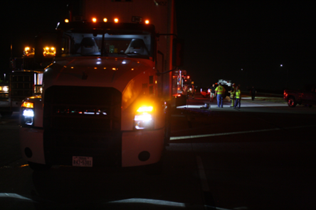 Nighttime scene of a large semi-truck with its headlights on and flashing lights on top, blocking part of the road, with construction workers in reflective vests working in the background.