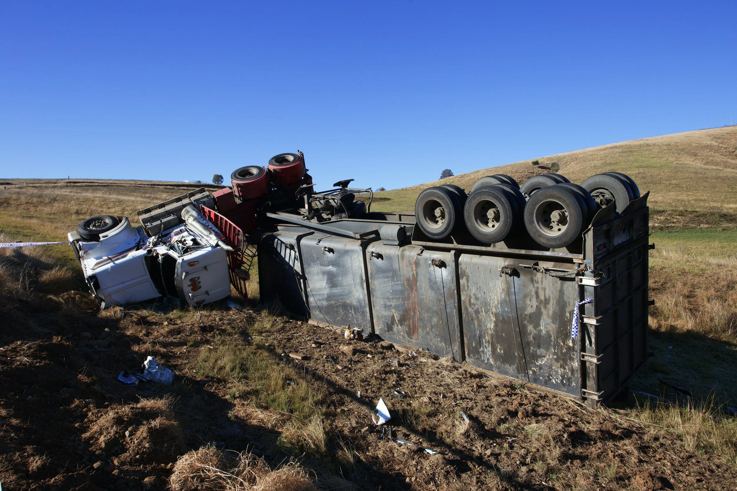 An overturned truck lying on its side in a grassy field under a clear blue sky.