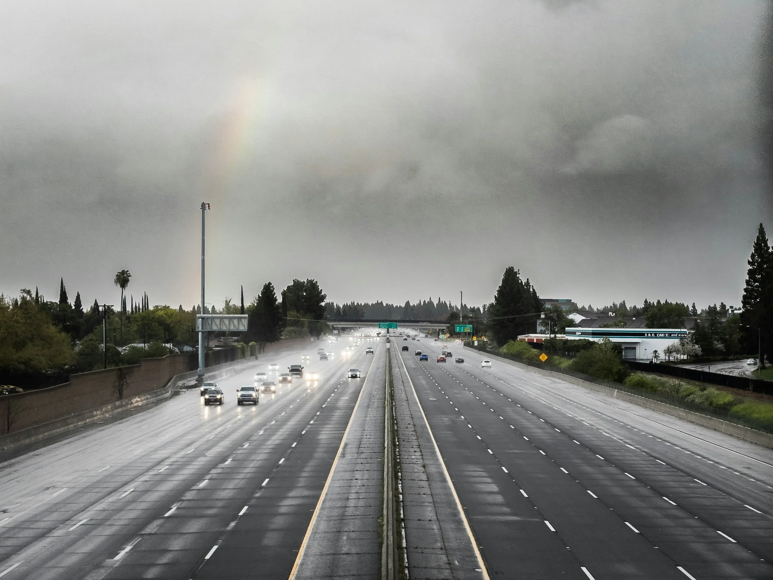 Empty wet highway with cars driving and rain overcast sky with rainbow in the background.