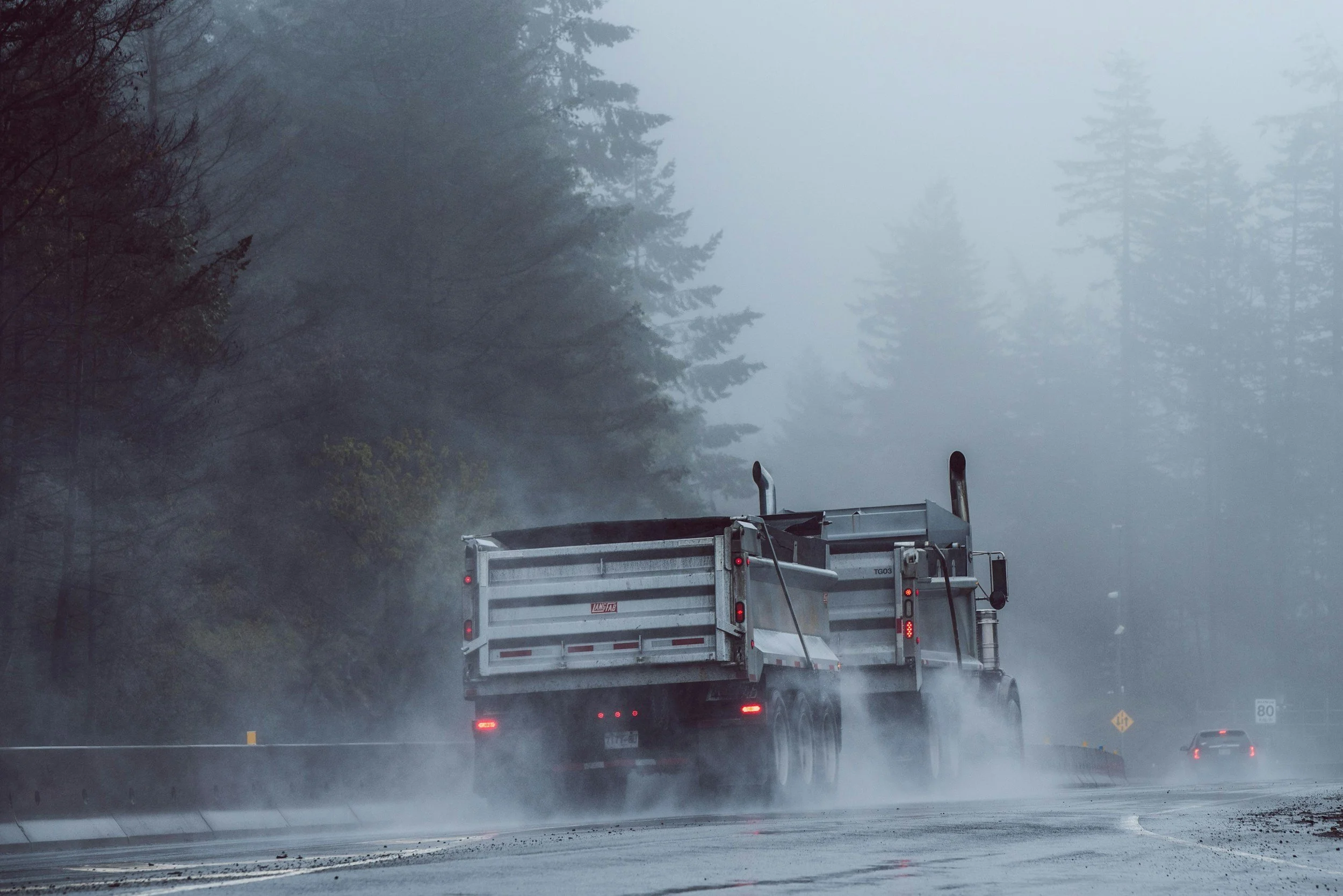 A large truck driving on a foggy wet road surrounded by trees.