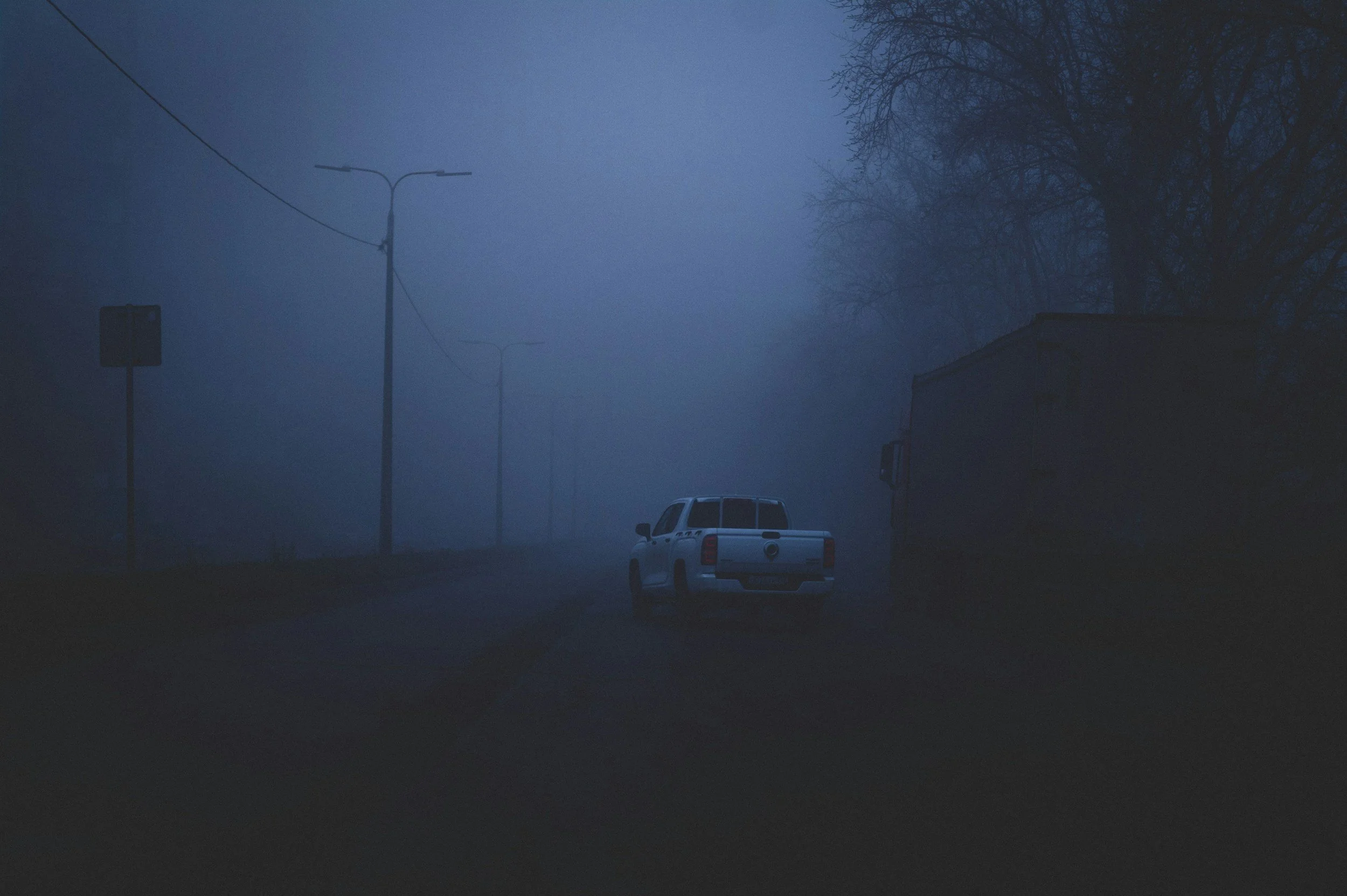 A white pickup truck and a large vehicle parked on a foggy, dark street at dusk or dawn, with streetlights and trees visible in the background.