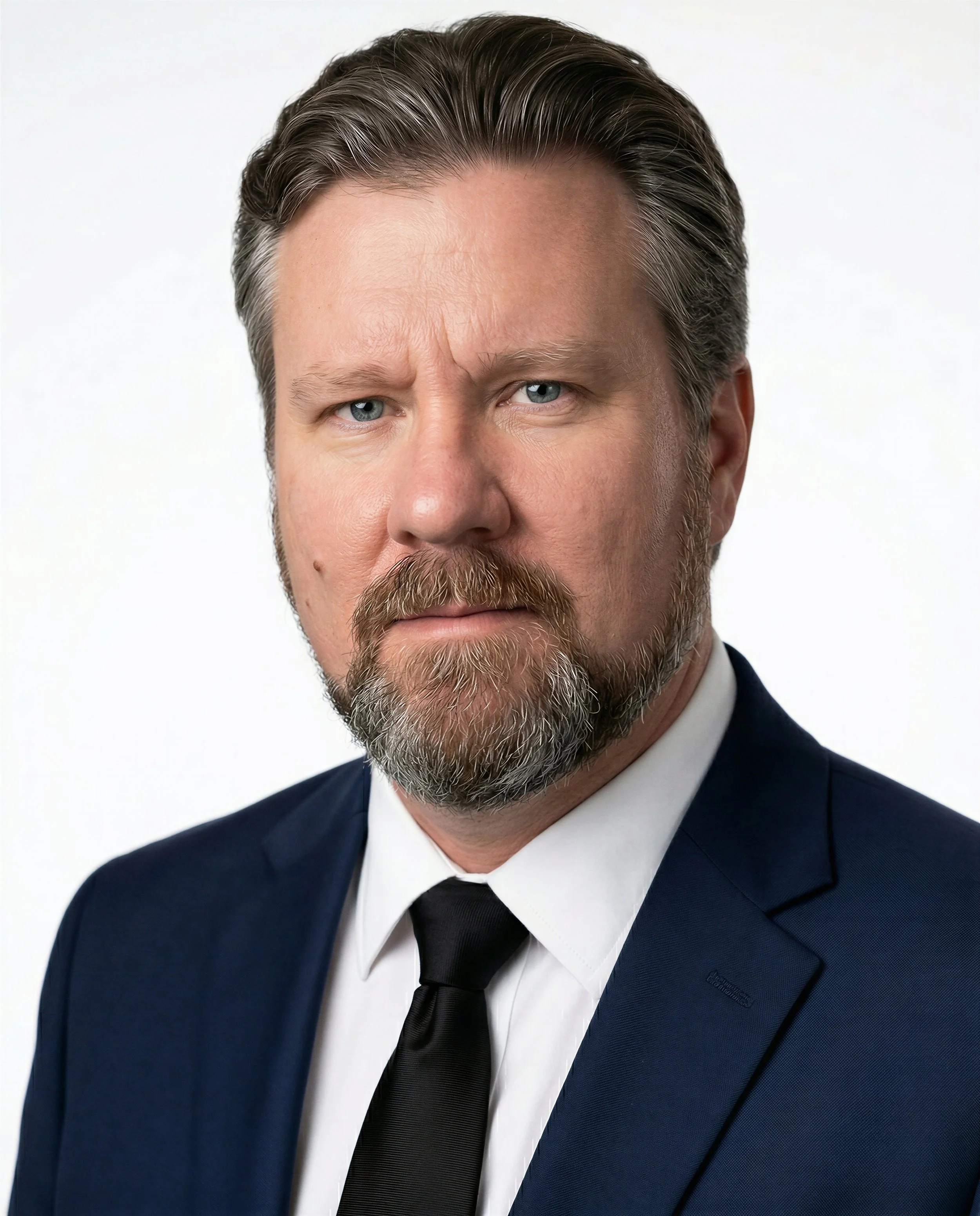 A middle-aged man with light skin, a full beard, and styled gray hair, wearing a dark suit, white shirt, and a solid tie, posing against a white background.