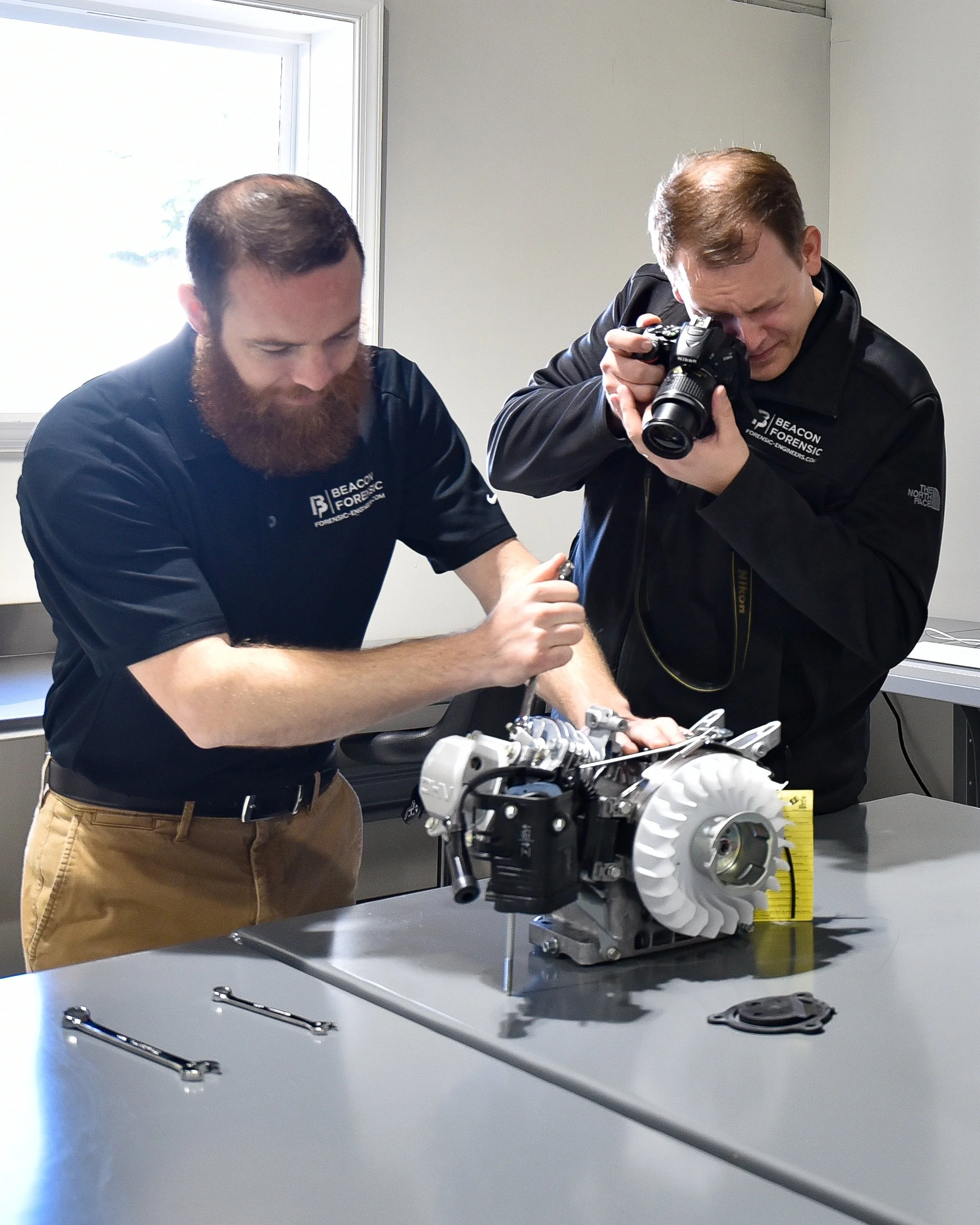 Two forensic engineers inspecting a pump motor in a laboratory, one holding a tool and the other using a camera.
