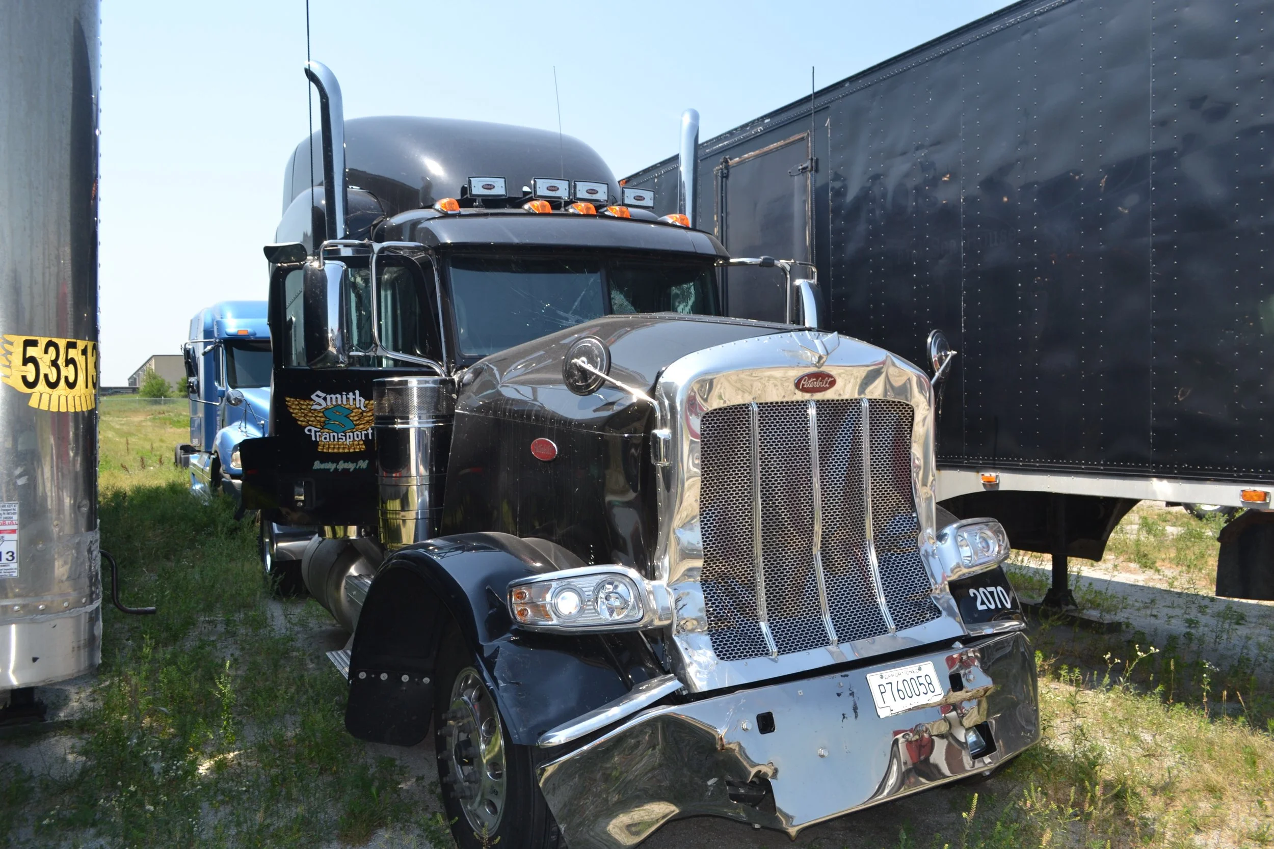 Damaged semi-truck with a crumpled front end parked between other trucks on a grassy lot.
