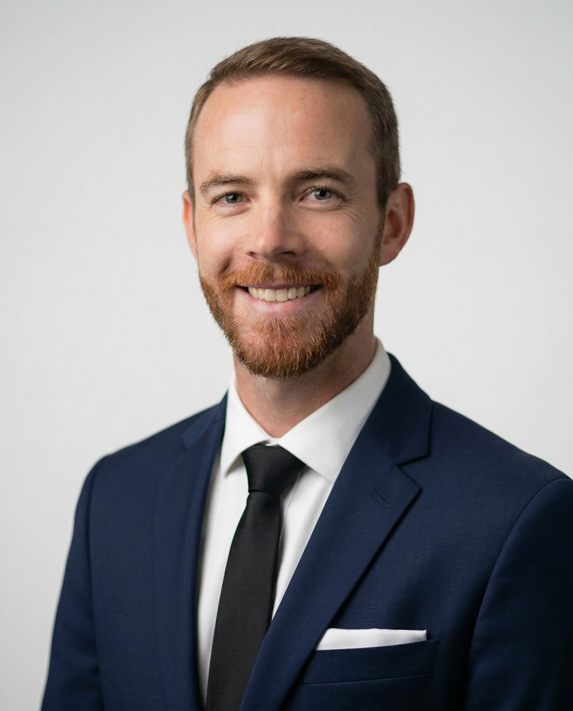 A portrait of a smiling man with a beard and brown hair, wearing a gray suit and a light blue tie, standing against a plain white wall.