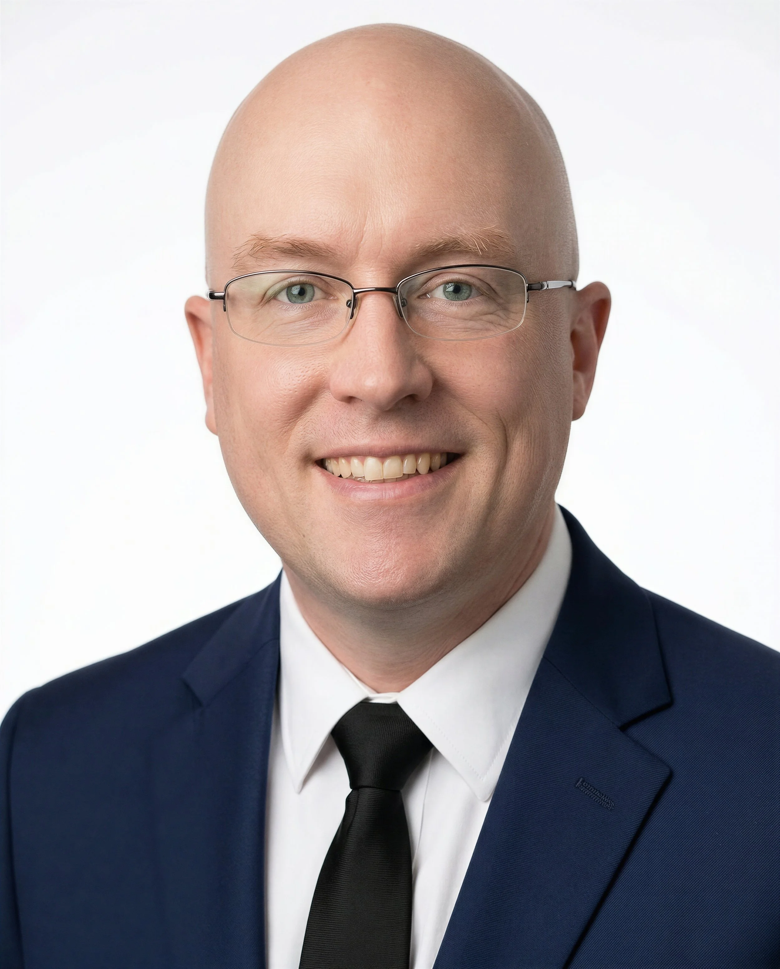 A man in a black suit, white shirt, and lack tie, wearing glasses, smiling at the camera against a plain, light-colored background.