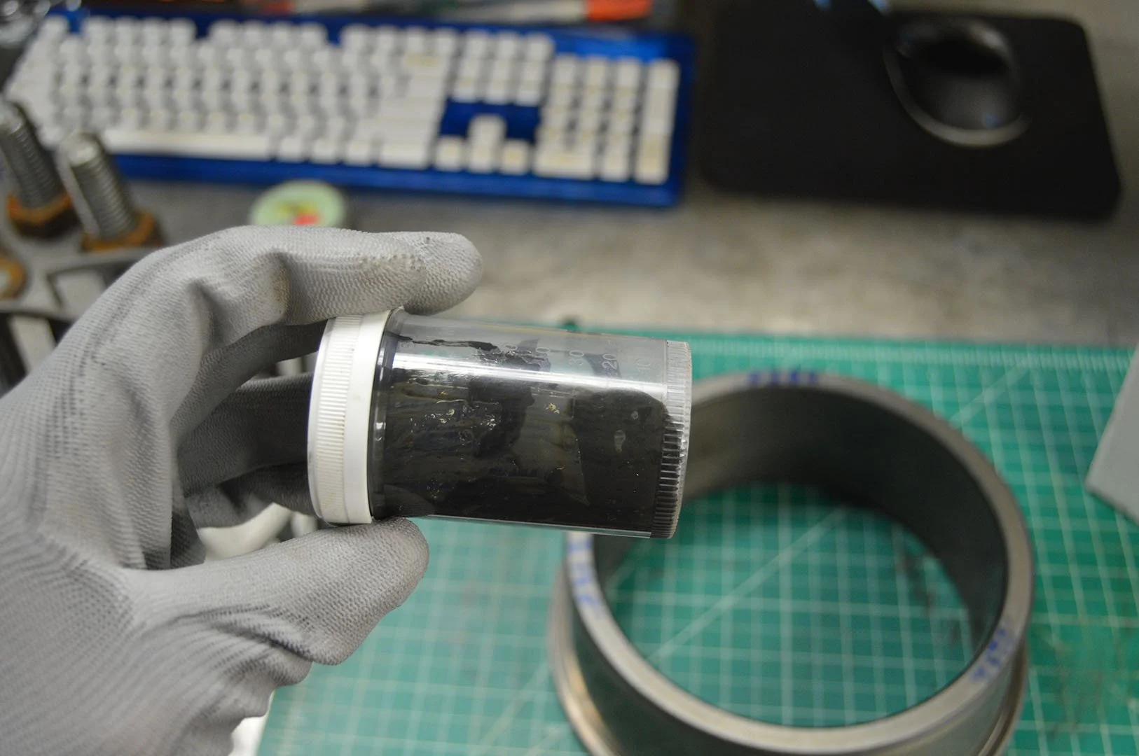 A gloved hand holding a small clear container filled with a black substance, with a black Z-shaped object inside, above a green cutting mat, with a metallic ring and various tools and objects in the background.