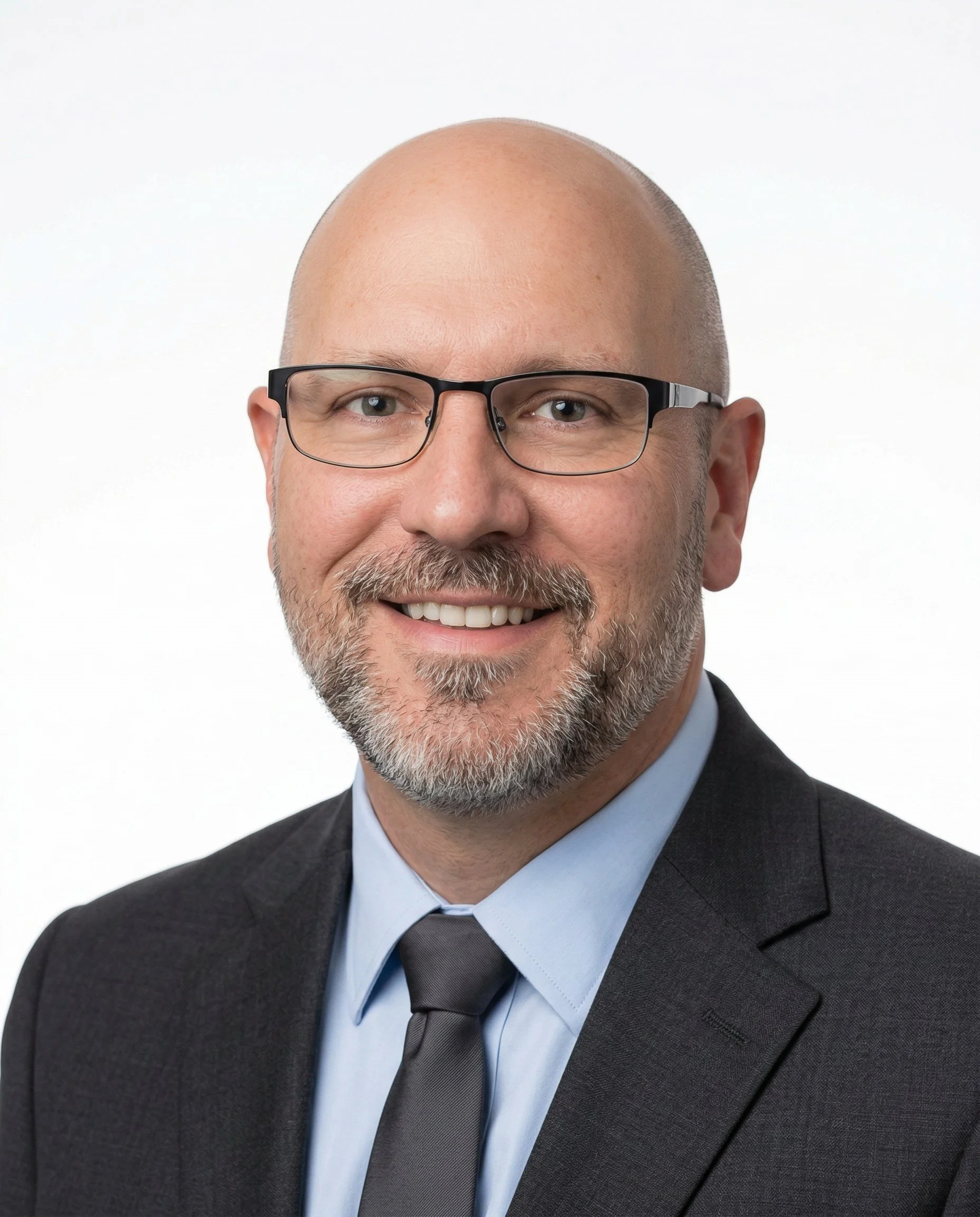 A smiling man with glasses, a beard, and a mustache, wearing a dark suit, light blue shirt, and a grey tie, posed against a neutral background.