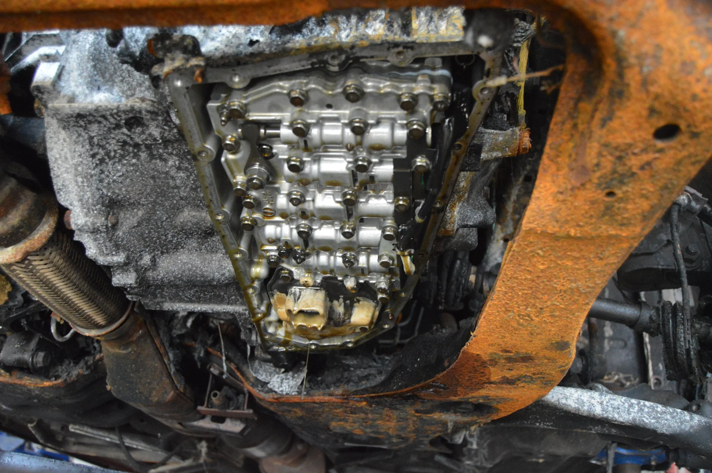 Under the car, exposed engine oil pan with oil and grime, rusty frame, and various mechanical components.