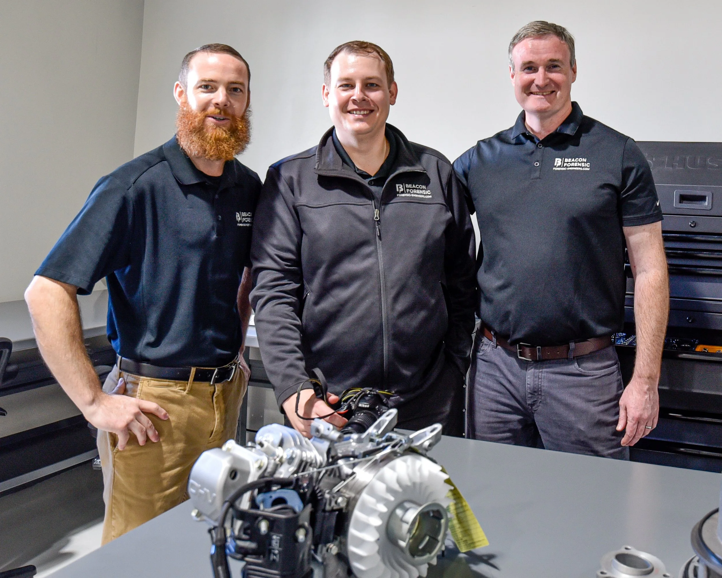 Three men standing in a room, smiling, with a small engine on a table in front of them. All three are wearing black or dark-colored shirts with the logo 'Beacon Forensic' on them.