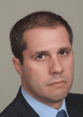 A man with short dark hair, wearing a dark suit and a light blue shirt, looking serious against a plain gray background.