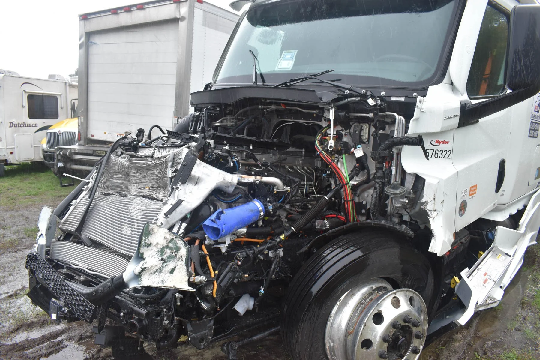 Damaged semi-truck with its front parts removed, exposing the engine and internal components, with damaged front bumper and fender. The truck is parked outdoors on a muddy ground, with other vehicles in the background.