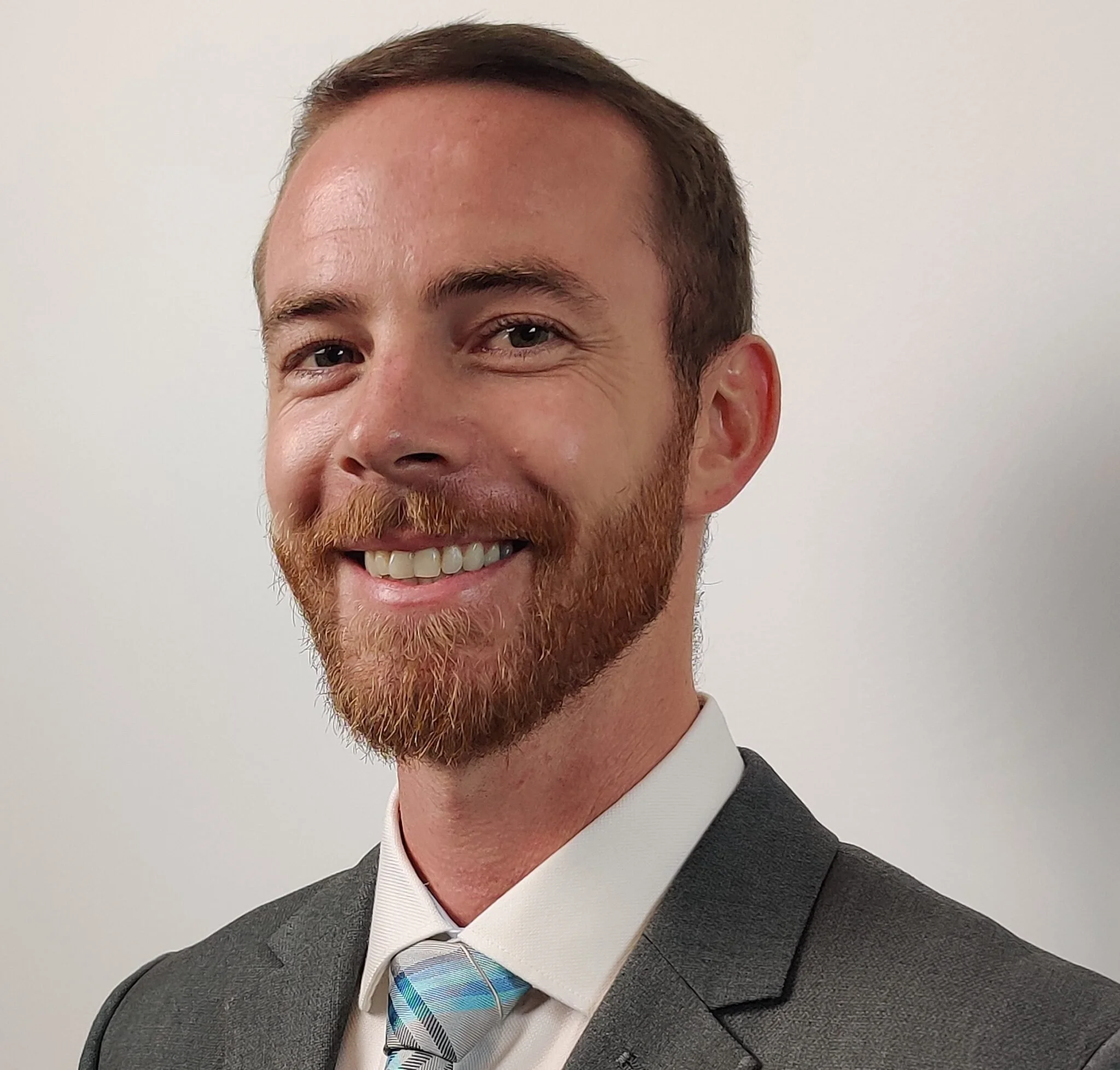 A professional man with a red beard, wearing a gray suit, white shirt, and a striped tie, smiling against a plain white background.