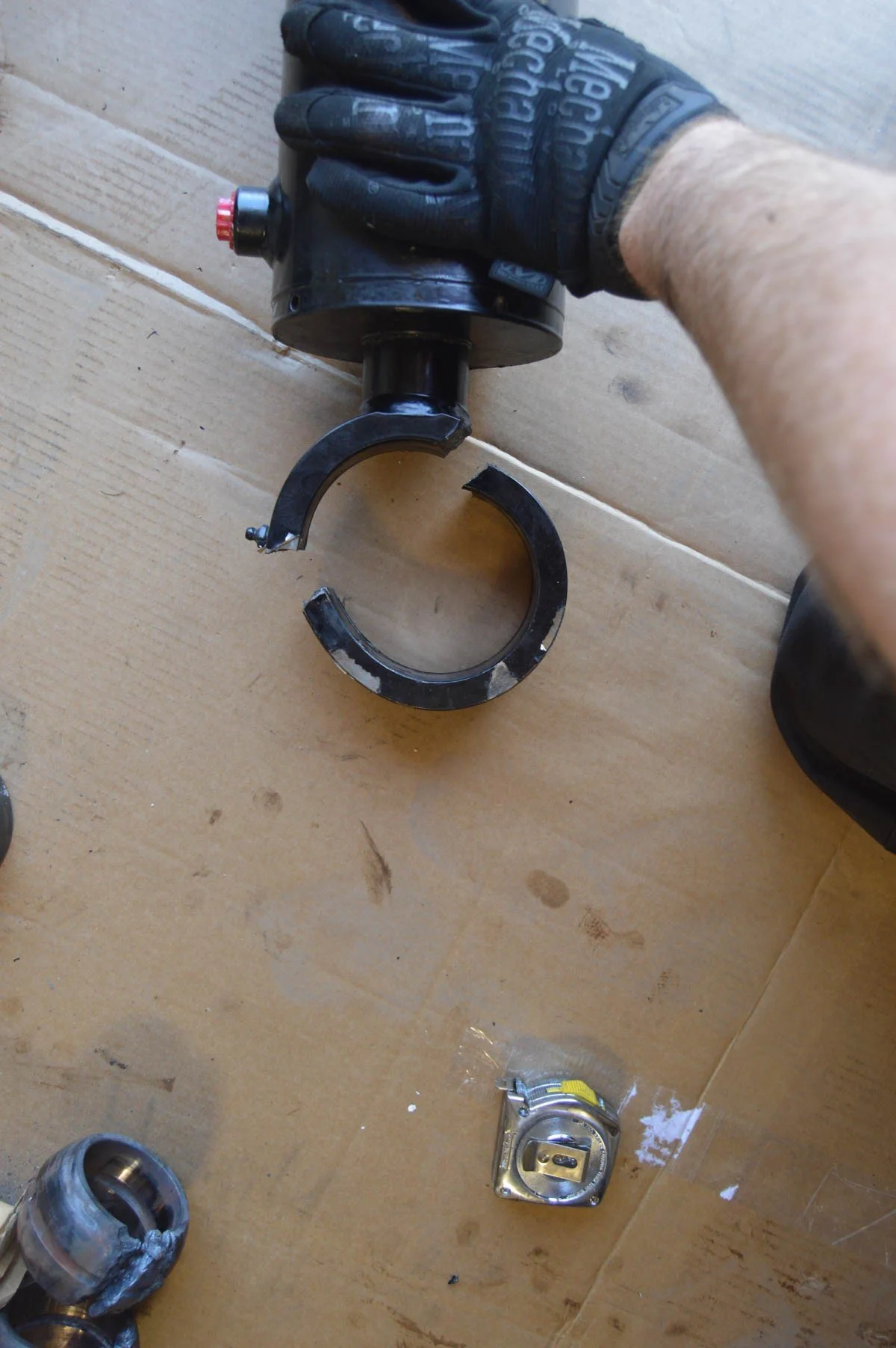 A person wearing black gloves holding a large black metal component with a clamp on the end, positioned over a wooden surface. There is a metallic electrical outlet on the wall below the component, and partially visible mechanical parts and tools.
