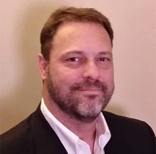 Portrait of a man with light skin, short brown hair, and a beard. He is wearing a black suit and white shirt, smiling slightly, against a plain beige wall background.