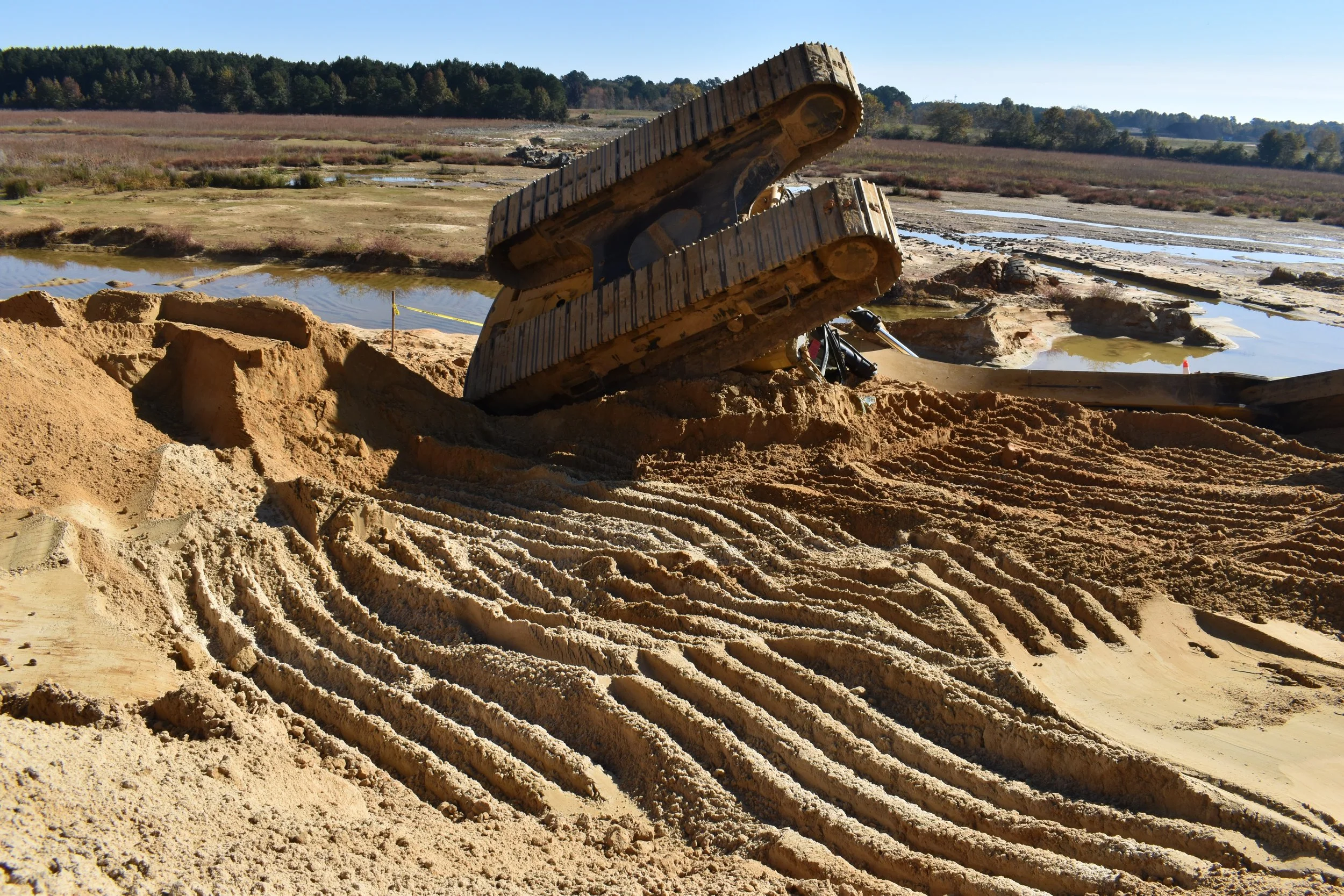 Construction site with excavator tracks in sand and muddy terrain, water pools and a distant tree line under a clear sky.
