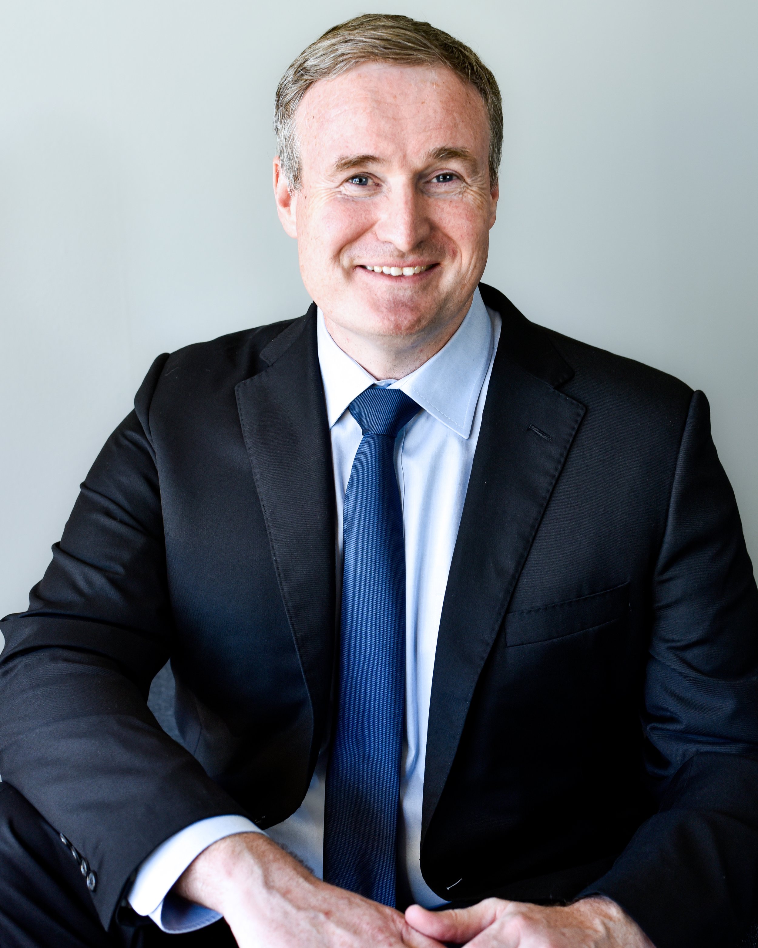 Professional man in a suit smiling with hands clasped, wearing a navy tie and white shirt, against a plain background.