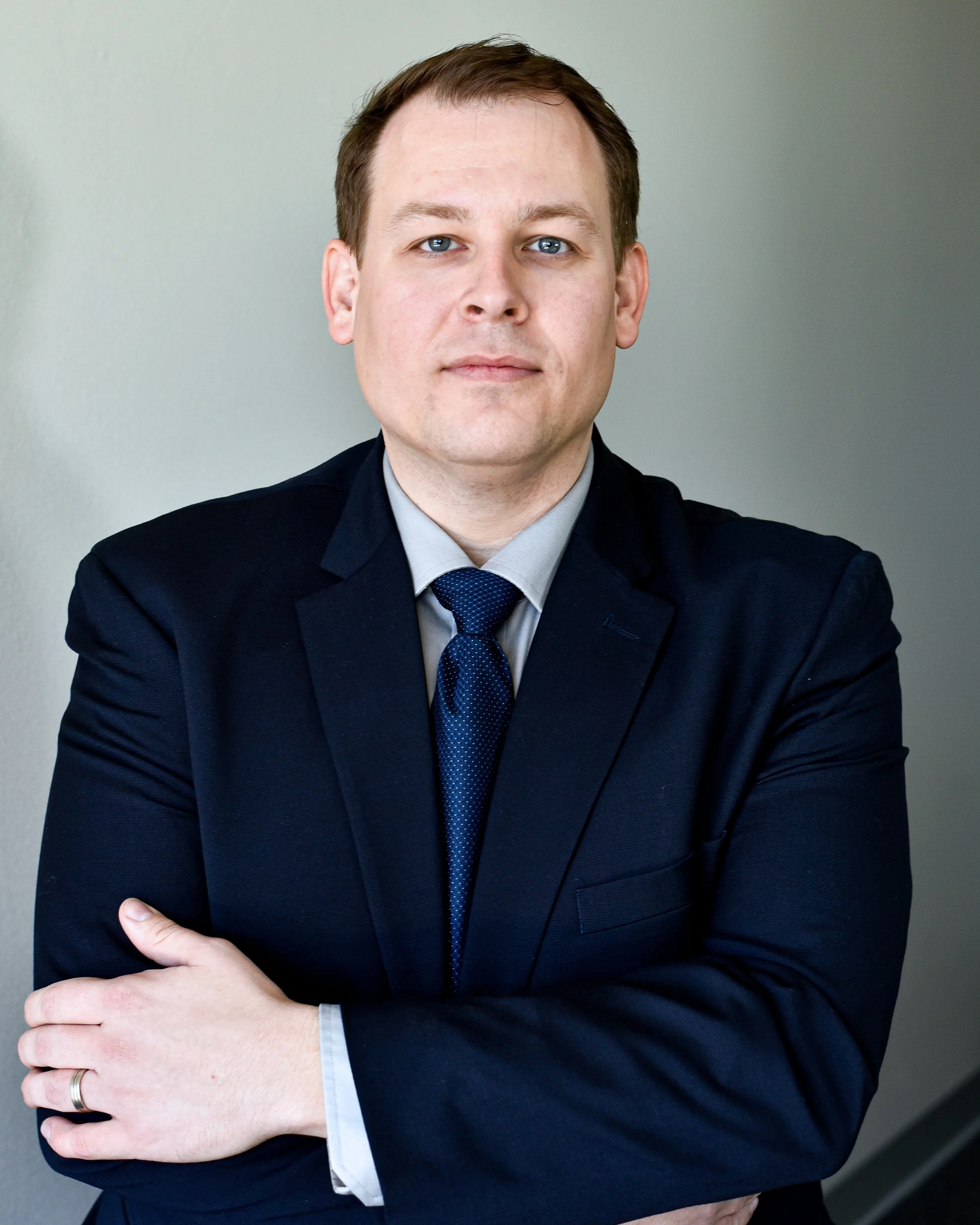 A professional man in a dark suit with crossed arms, wearing a light shirt and a blue tie, standing against a plain background.