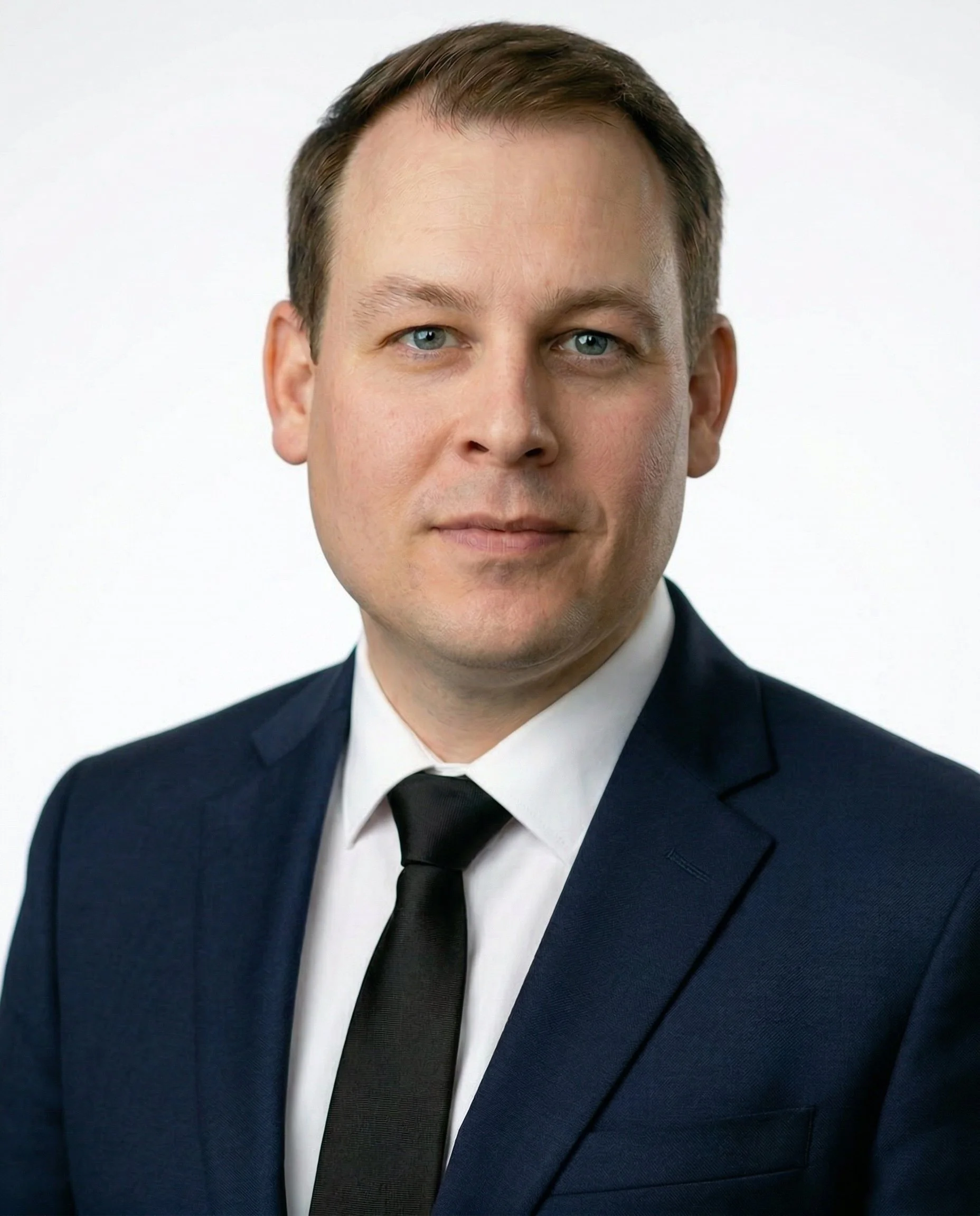 A man in a dark suit with a light-colored shirt and dark tie, standing with folded arms against a plain wall.