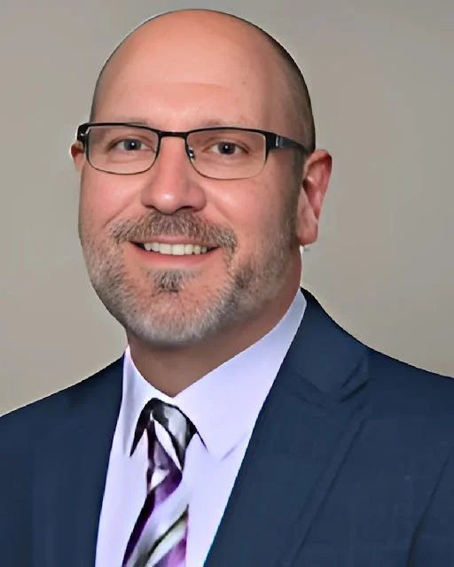 A smiling man with glasses, a beard, and a mustache, wearing a dark suit, light blue shirt, and a striped tie, posed against a neutral background.