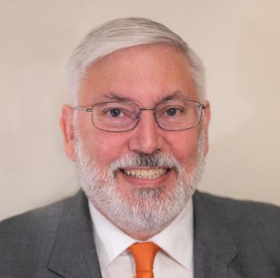 A smiling older man with gray hair, a beard, and glasses wearing a gray suit, white shirt, and orange tie.