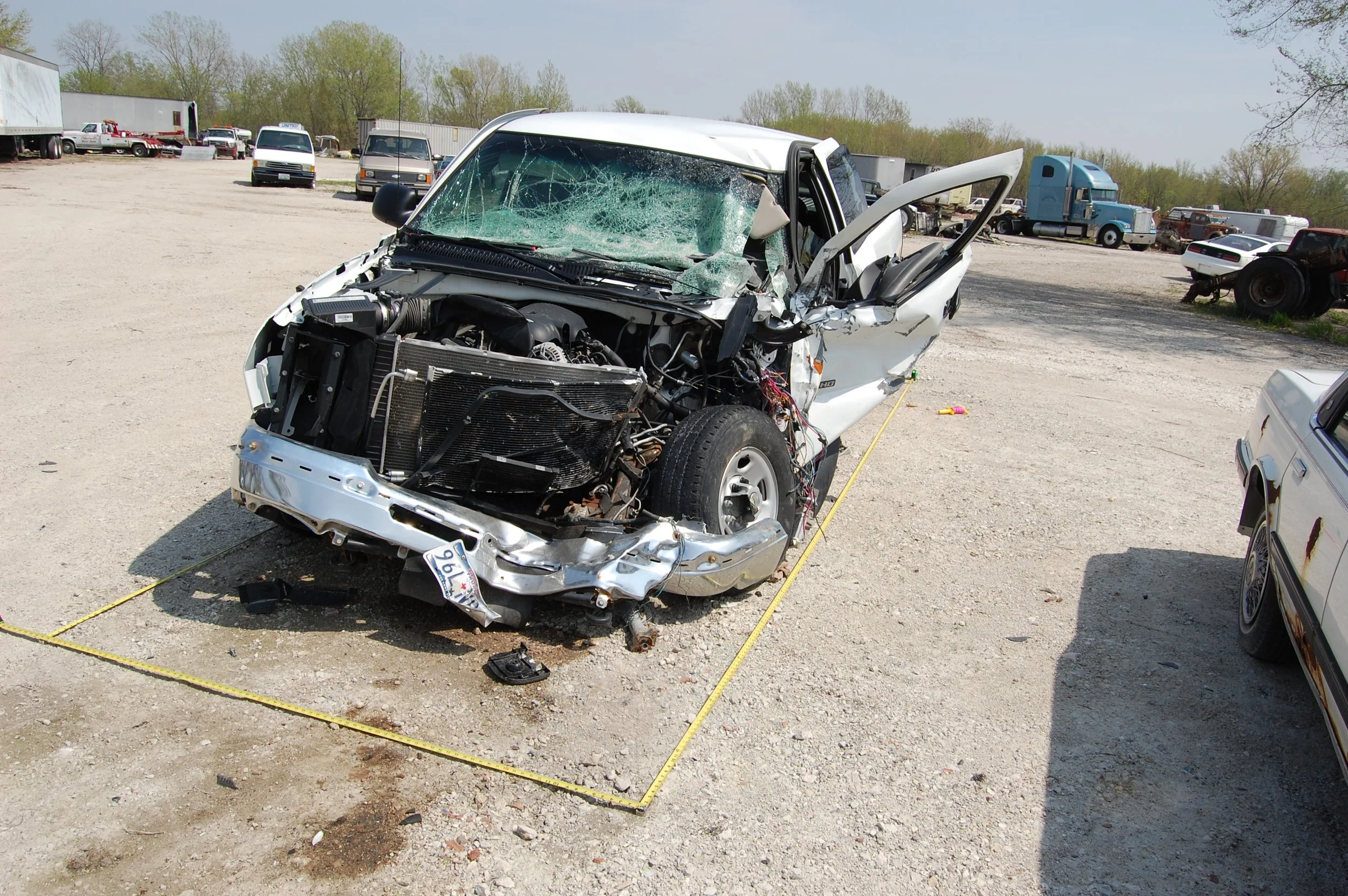 A white car with severe front-end damage and shattered windshield in a salvage yard.
