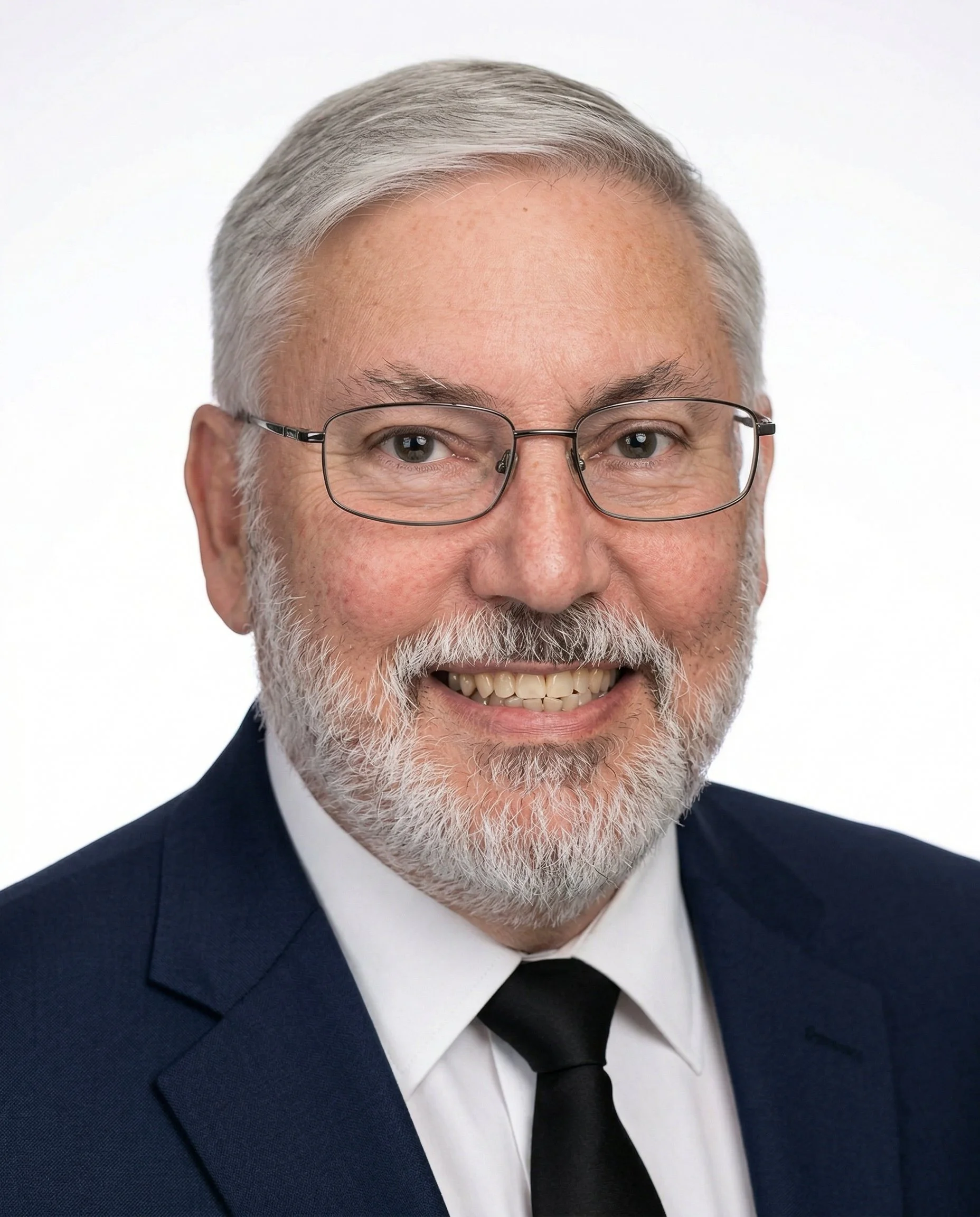 Portrait of a man with a red beard and wearing a navy blue suit, white shirt, and a plaid tie, sitting with arms crossed on a gray chair against a plain light-colored background.