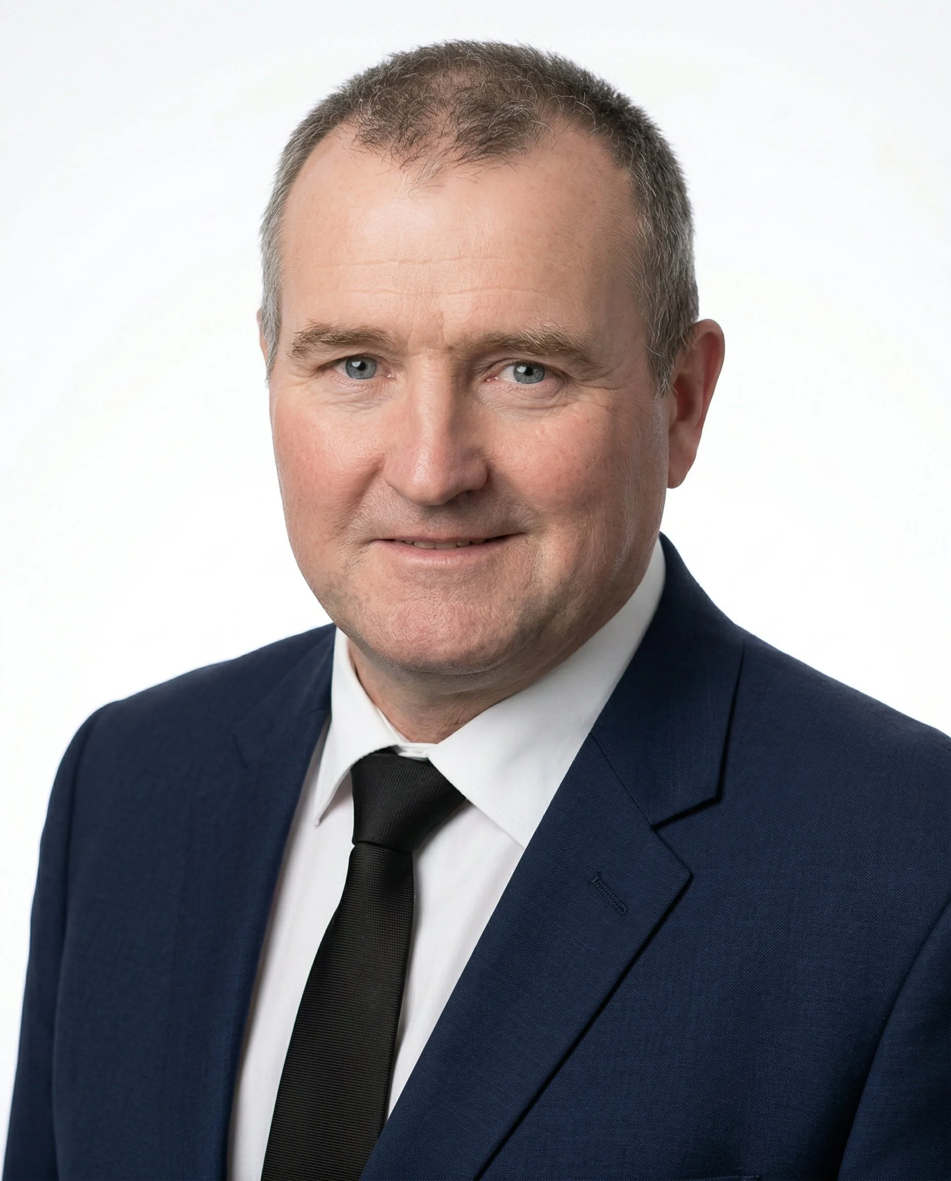 A man wearing a dark suit, white shirt, and black solid tie, smiling at the camera against a light gray background.