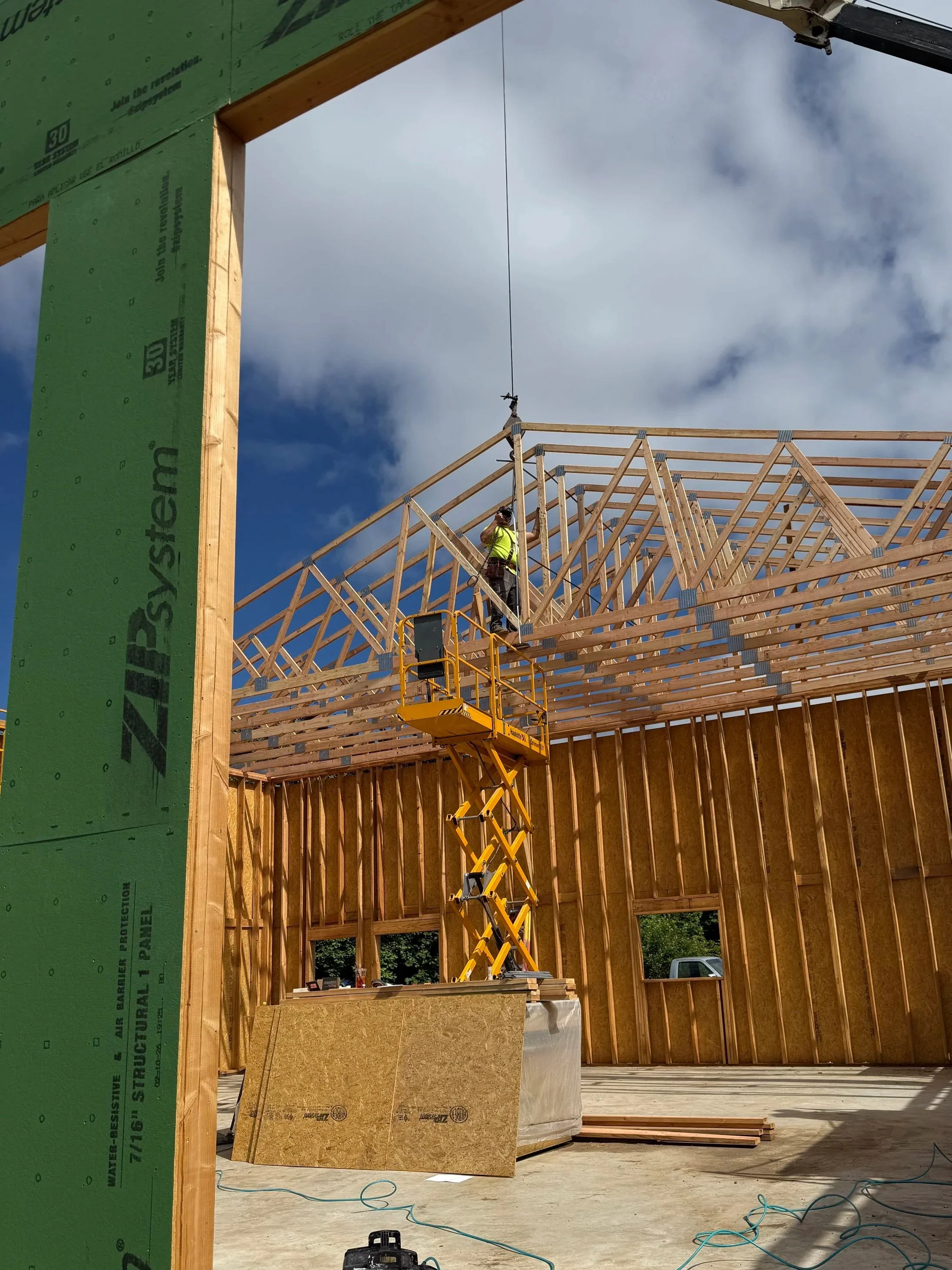 Construction site with wooden framing for a building, worker on a yellow lift working near the roof trusses, cloudy sky in the background.