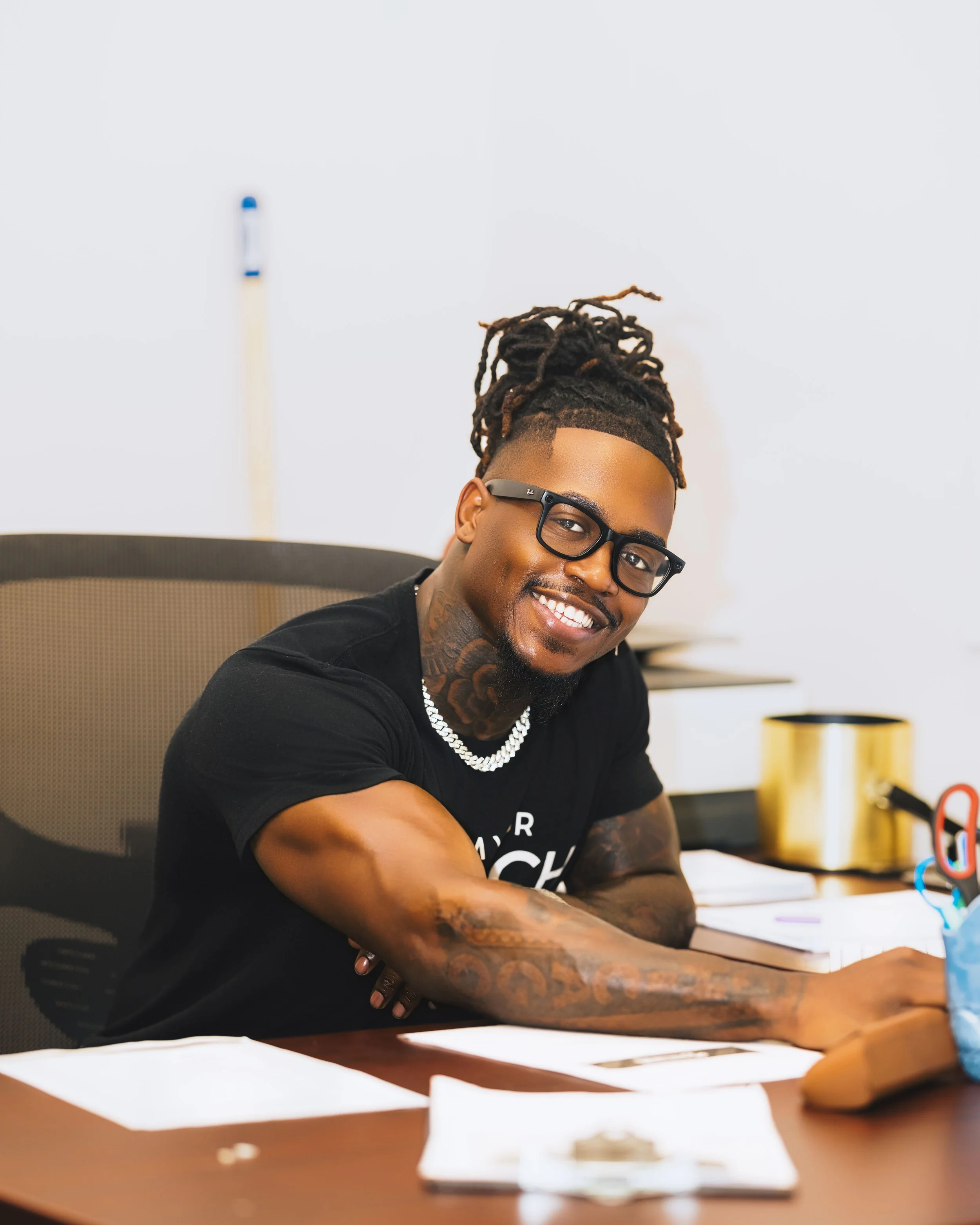 Smiling man with glasses and tattoos seated at a desk in an office, with papers, a pen holder, and a gold-colored object on the desk.