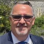 A smiling man wearing sunglasses, a blue suit, and a tie outdoors with greenery and a brick building in the background.