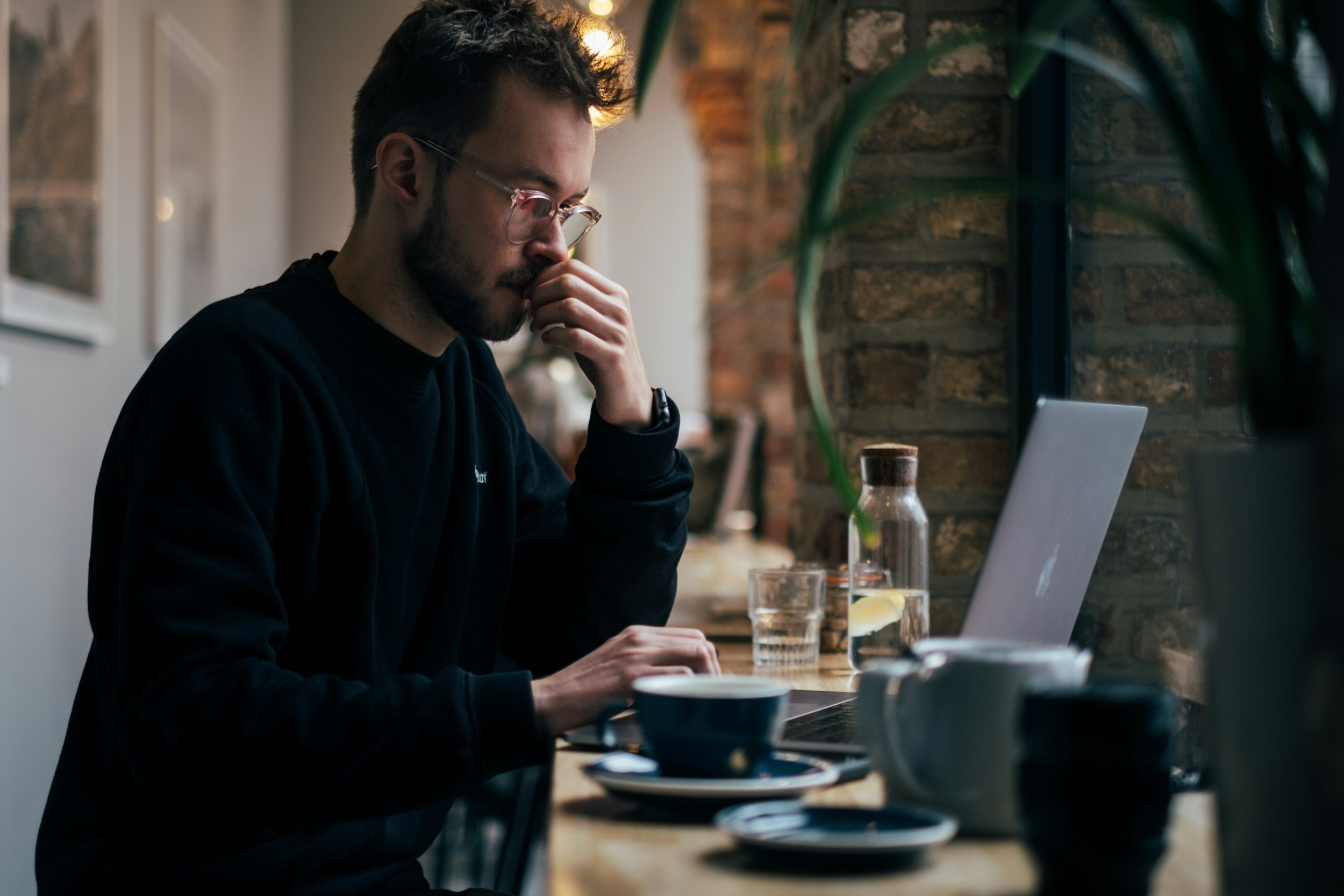 Person in glasses using a laptop in a cafe, appearing thoughtful. Coffee cups and a glass pitcher in foreground, exposed brick wall in background.