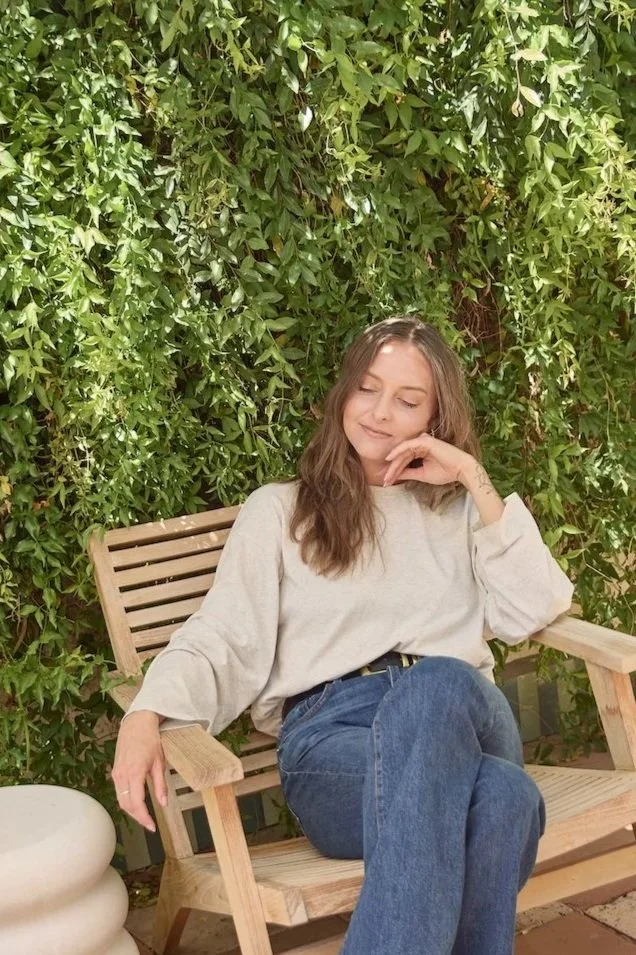 A woman sitting on a wooden bench in front of a wall of green ivy leaves, with her eyes closed and a content smile.