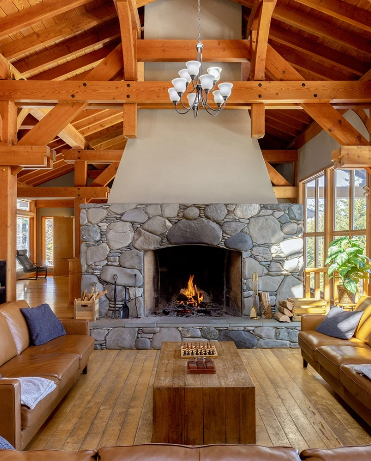 Living room with a stone fireplace, wooden ceiling beams, and leather sofas. A chessboard is on a wooden coffee table, and the room has large windows with a view of trees outside.