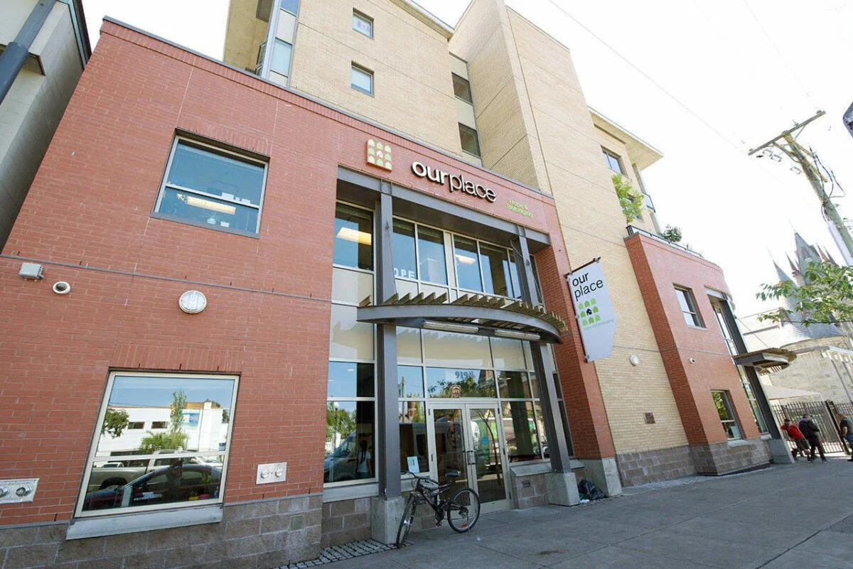 Exterior view of a modern multi-story building with a red brick facade and a sign that reads 'ourplace.' There are windows, a bicycle parked at the entrance, and two people walking on the sidewalk.