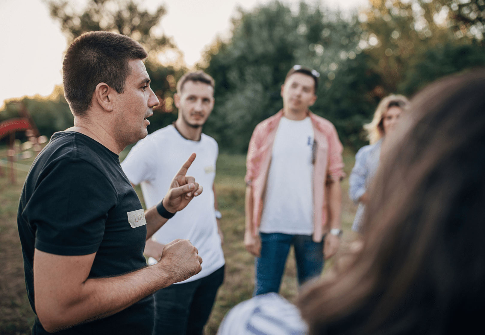 Man speaking to a group of four people outdoors during the daytime, with trees in the background.