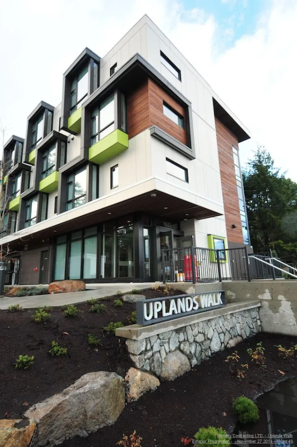 Modern multi-story apartment building with white, black, and wooden exterior, green balcony accents, and a sign that reads 'UPLANDS WALK' in front of landscaped area. Affordable Housing
