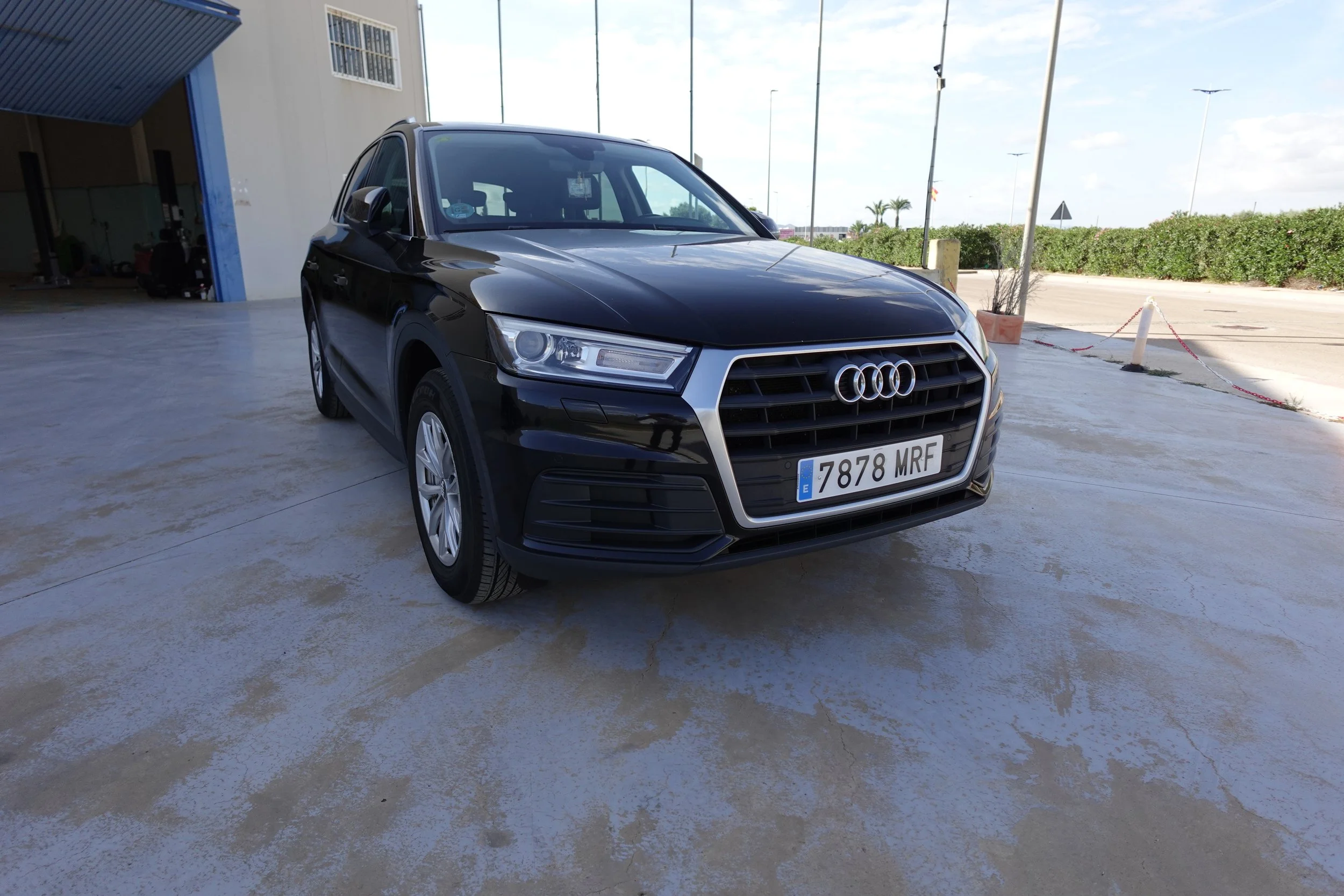 A black Audi SUV parked outdoors on a concrete surface near a building, with a street and greenery visible in the background.