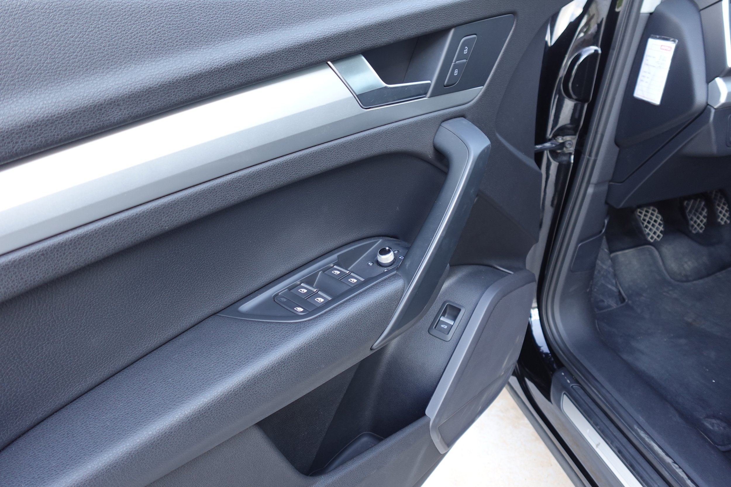 Inside view of a car's driver's side door with window controls, lock, and handle, with the car's interior and foot pedals visible.