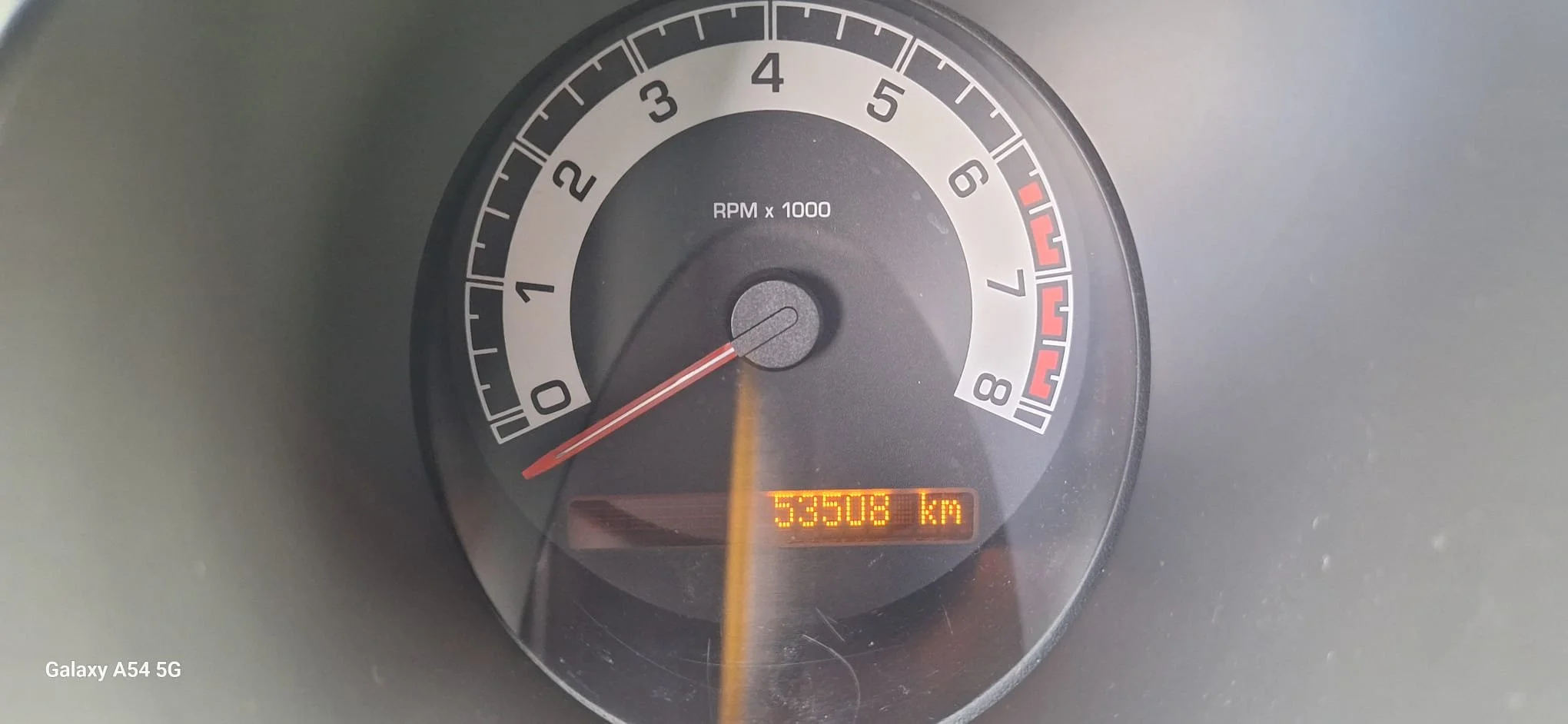 Inside a vehicle, view of the black steering wheel with a Renault logo, dashboard with speedometer, tachometer, fuel gauge, and a small digital display showing temperature and other indicators. To the right, part of the climate control system and center console are visible.