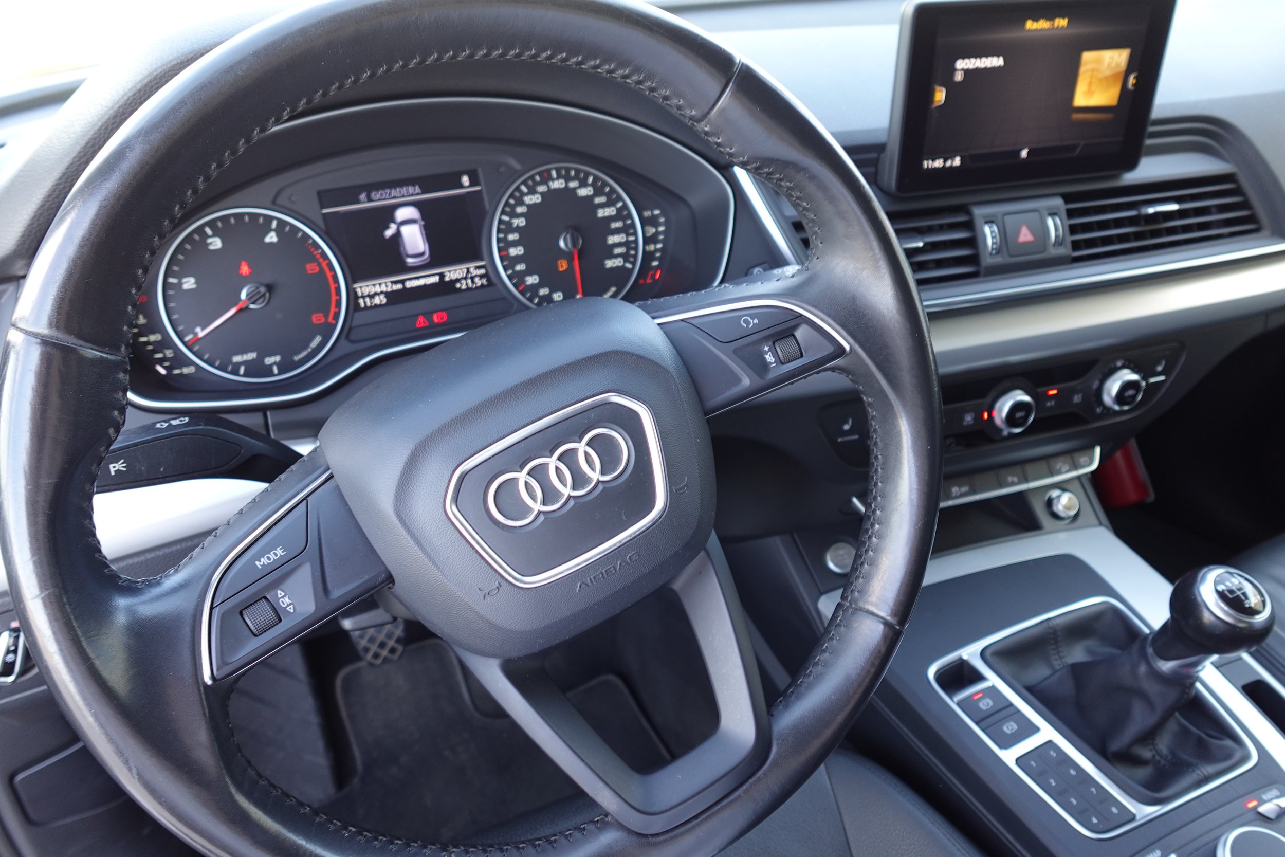Inside the driver's side of an Audi car, showing the steering wheel with Audi logo, dashboard with instrument cluster, digital display, and a infotainment screen mounted on the dashboard.