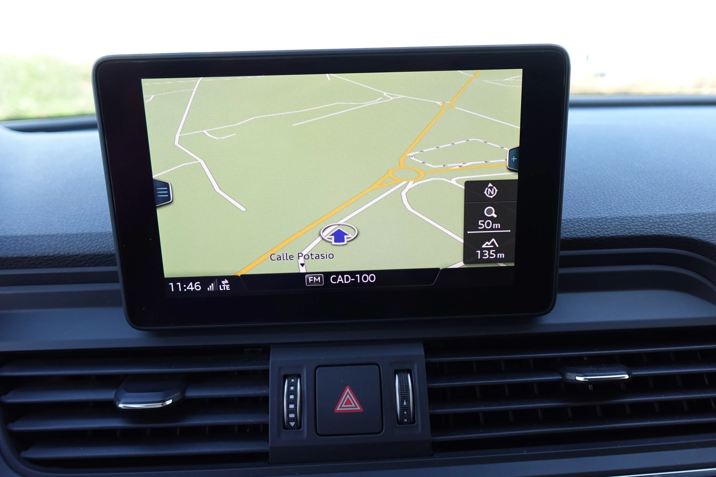 Car's dashboard GPS screen displaying a map with roads and a route, showing street name 'Calle Potasio', time 11:46, LTE signal, and radio station 'FM CAD-100'.
